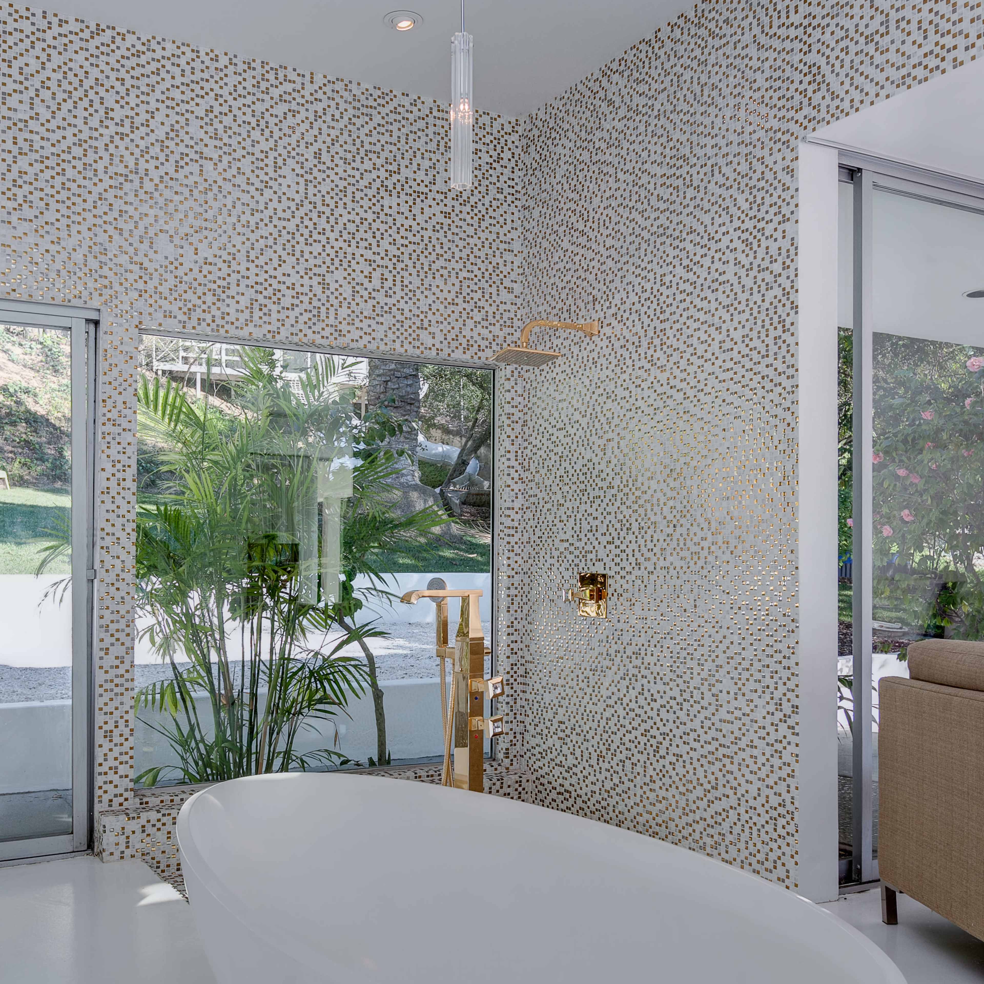 The bathroom features a freestanding white bathtub with a gold shower fixture against a mosaic tiled wall, surrounded by large glass windows and greenery.