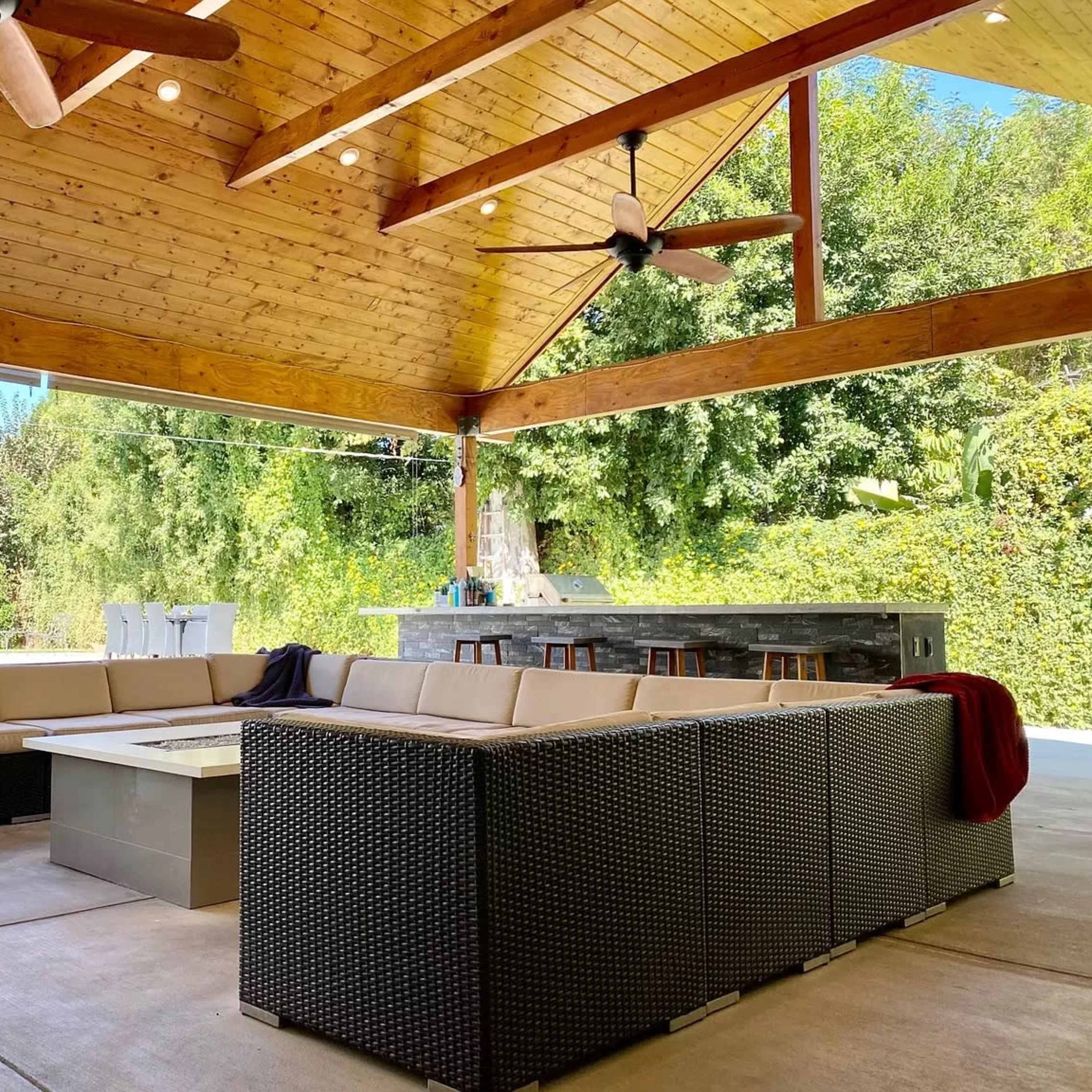 A covered outdoor seating area features a sectional sofa arranged around a central coffee table with a kitchen space in the background.
