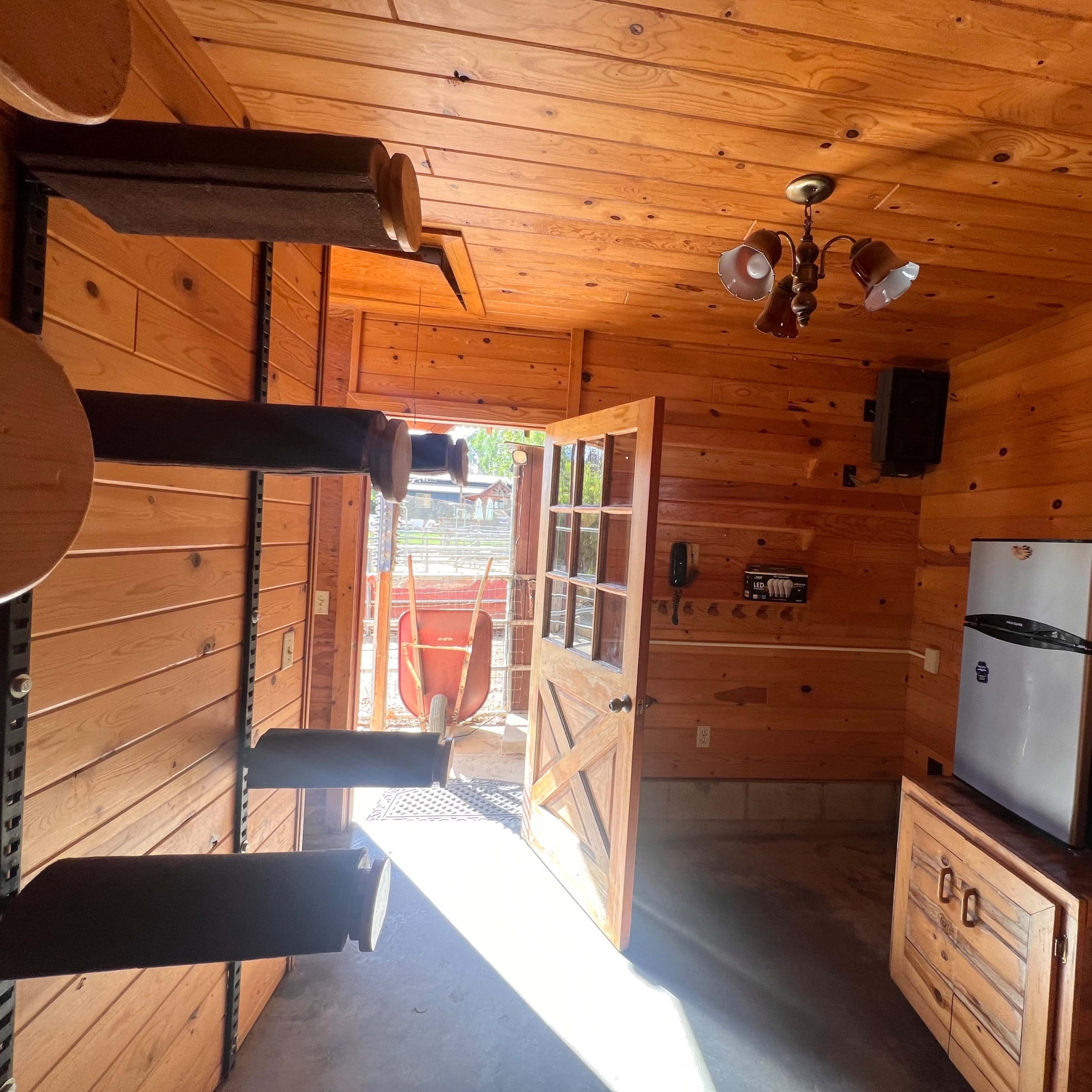 A wooden interior of a cabin with a door open to the outside, revealing a small fridge and shelves on one wall.