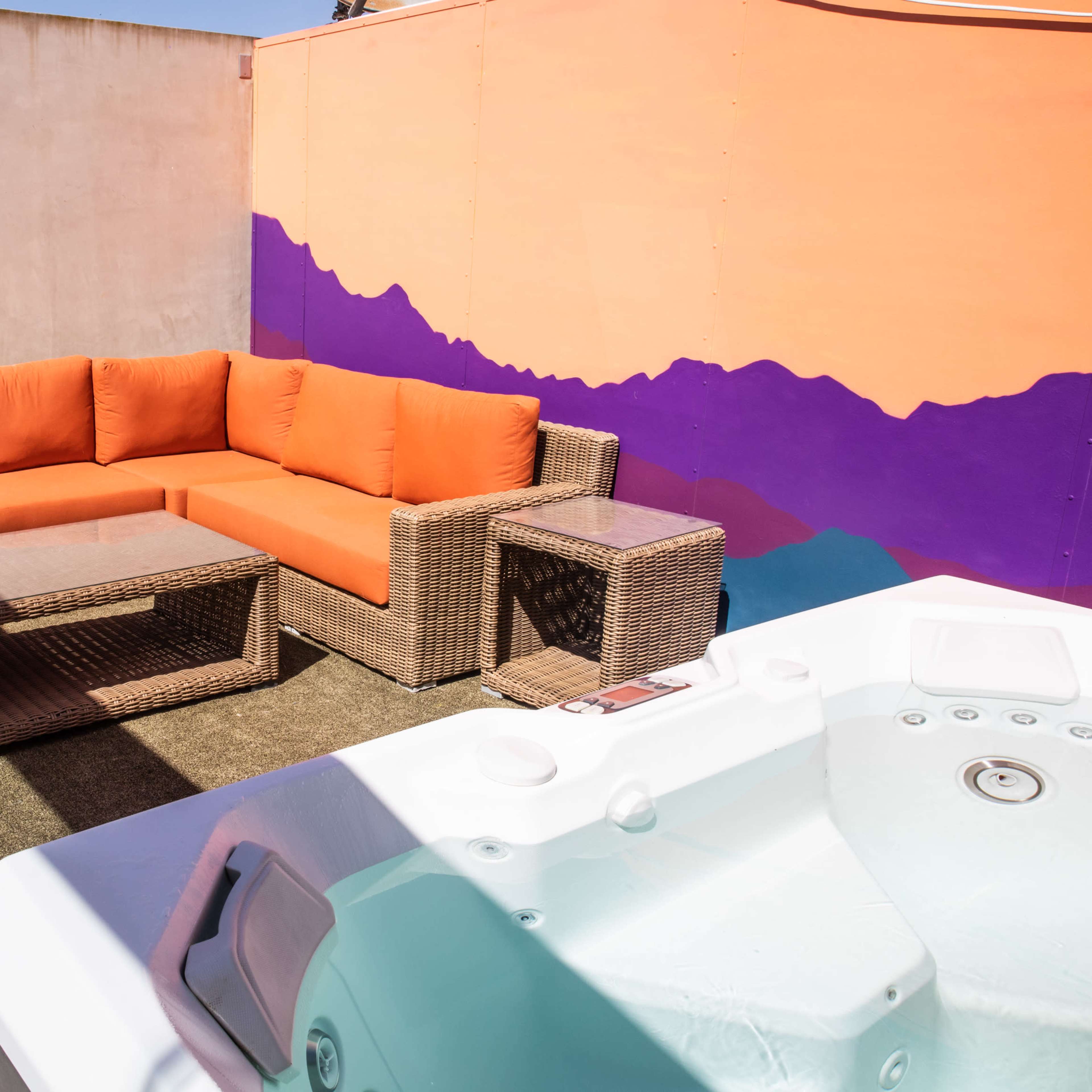 The scene features a rooftop patio with a large orange sectional sofa, a glass-top table, and a hot tub, all set against a colorful mural backdrop.