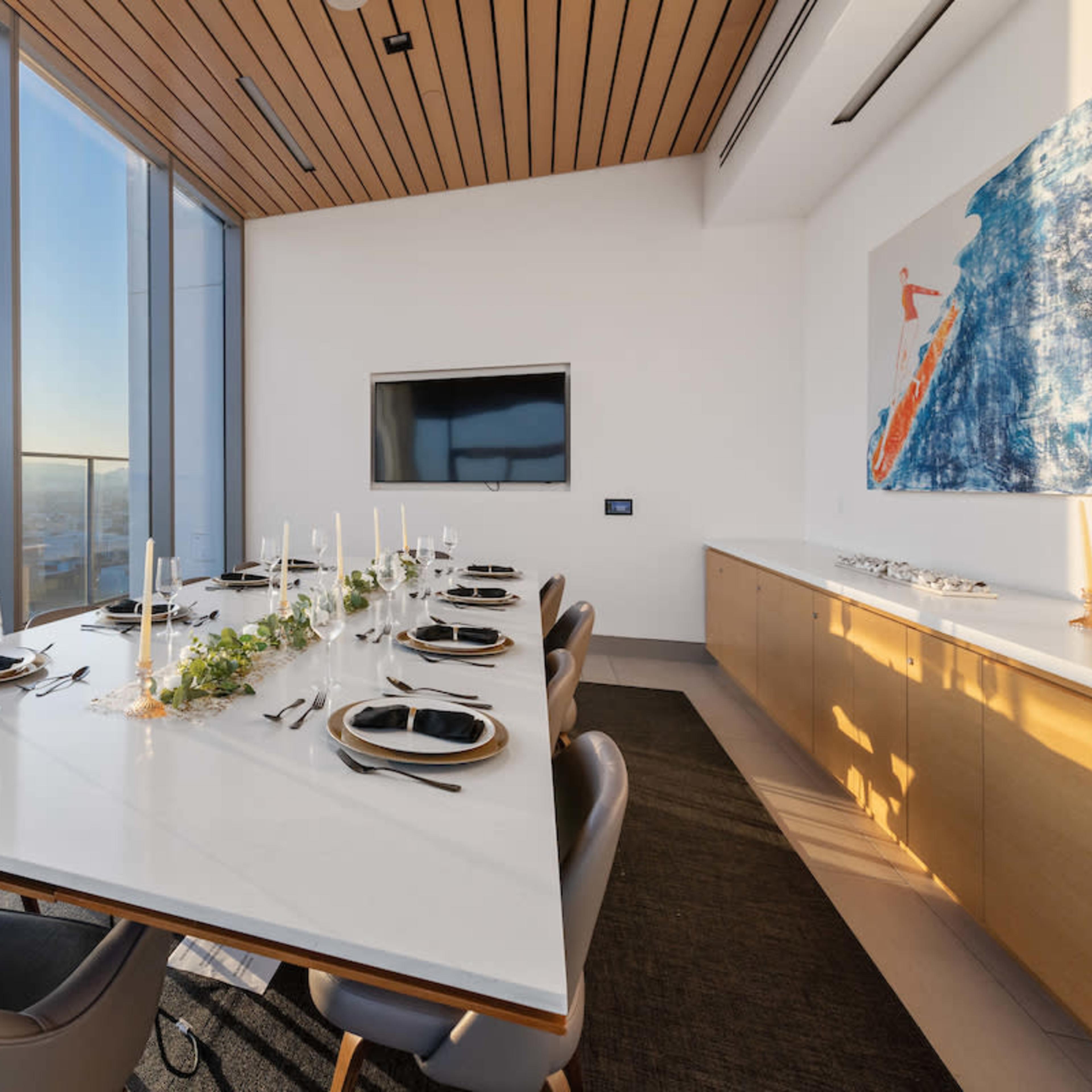 A modern dining room features a long table set for dinner, with large windows offering a view of the sunset.