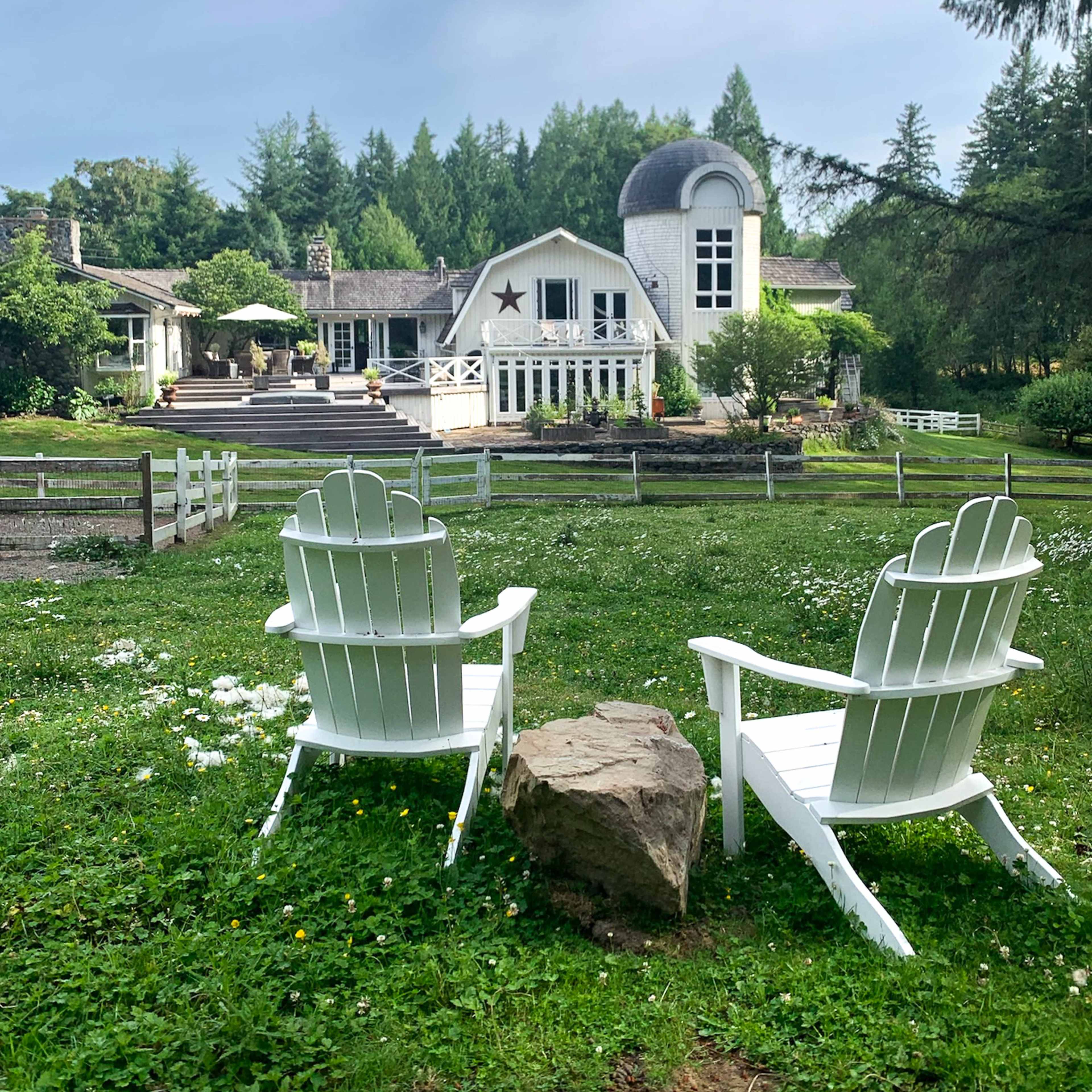 A pair of white Adirondack chairs sits on a grassy area in front of a house with a distinctive dome and star, surrounded by trees and gardens.