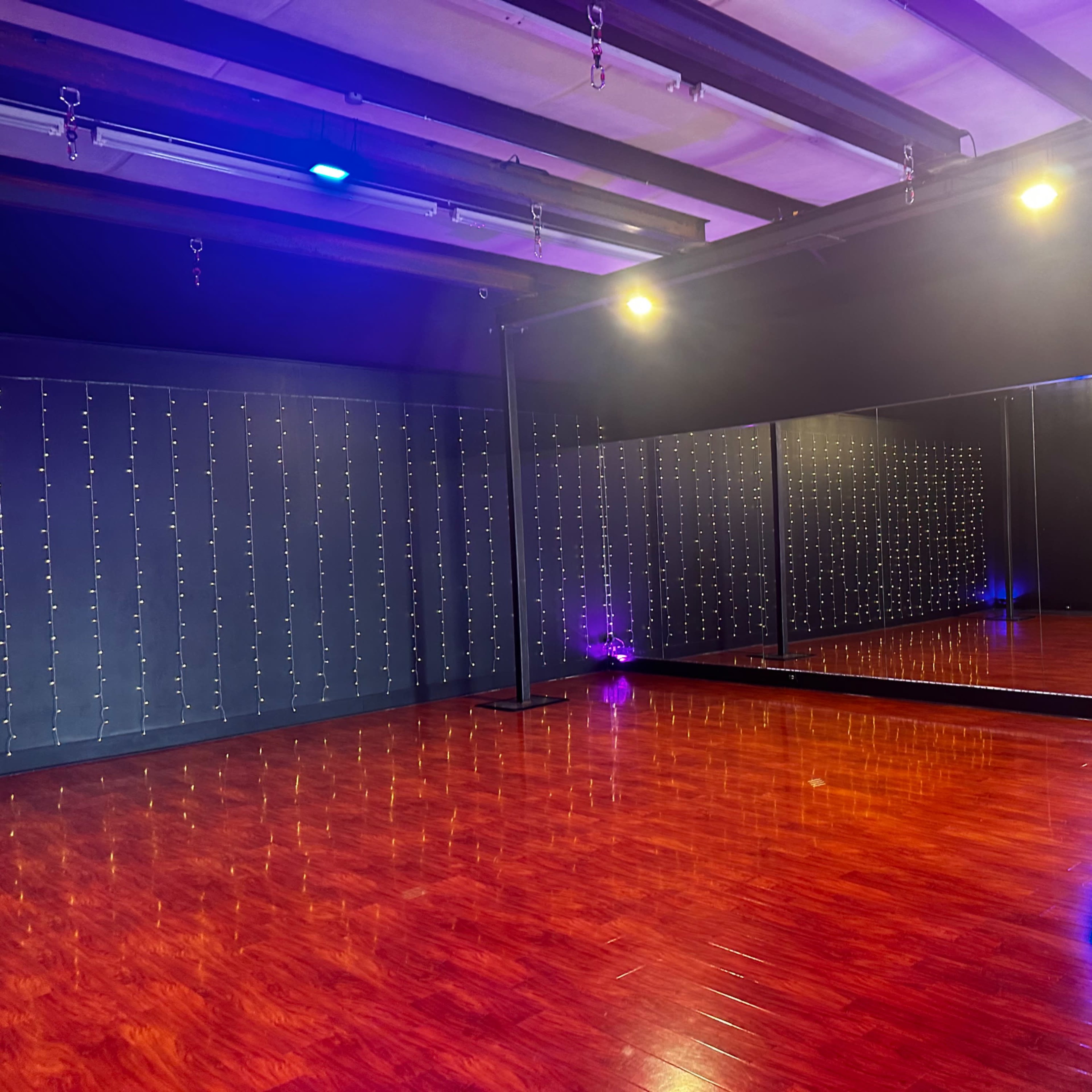 A spacious dance studio featuring hardwood floors, a large mirror, and walls adorned with string lights.