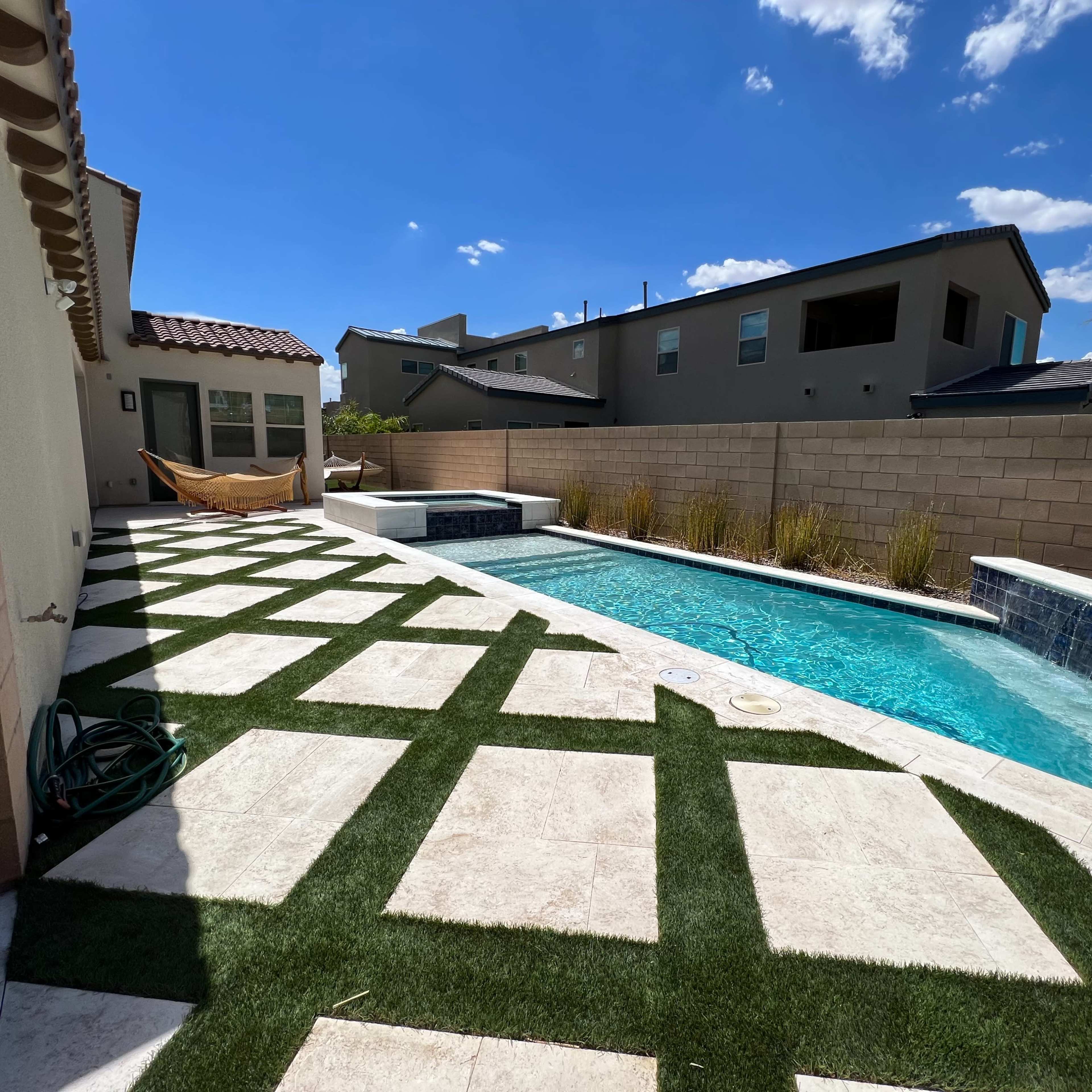 A residential backyard featuring a swimming pool, decorative landscaping with artificial grass, and a patio area surrounded by a stone wall and houses in the background.