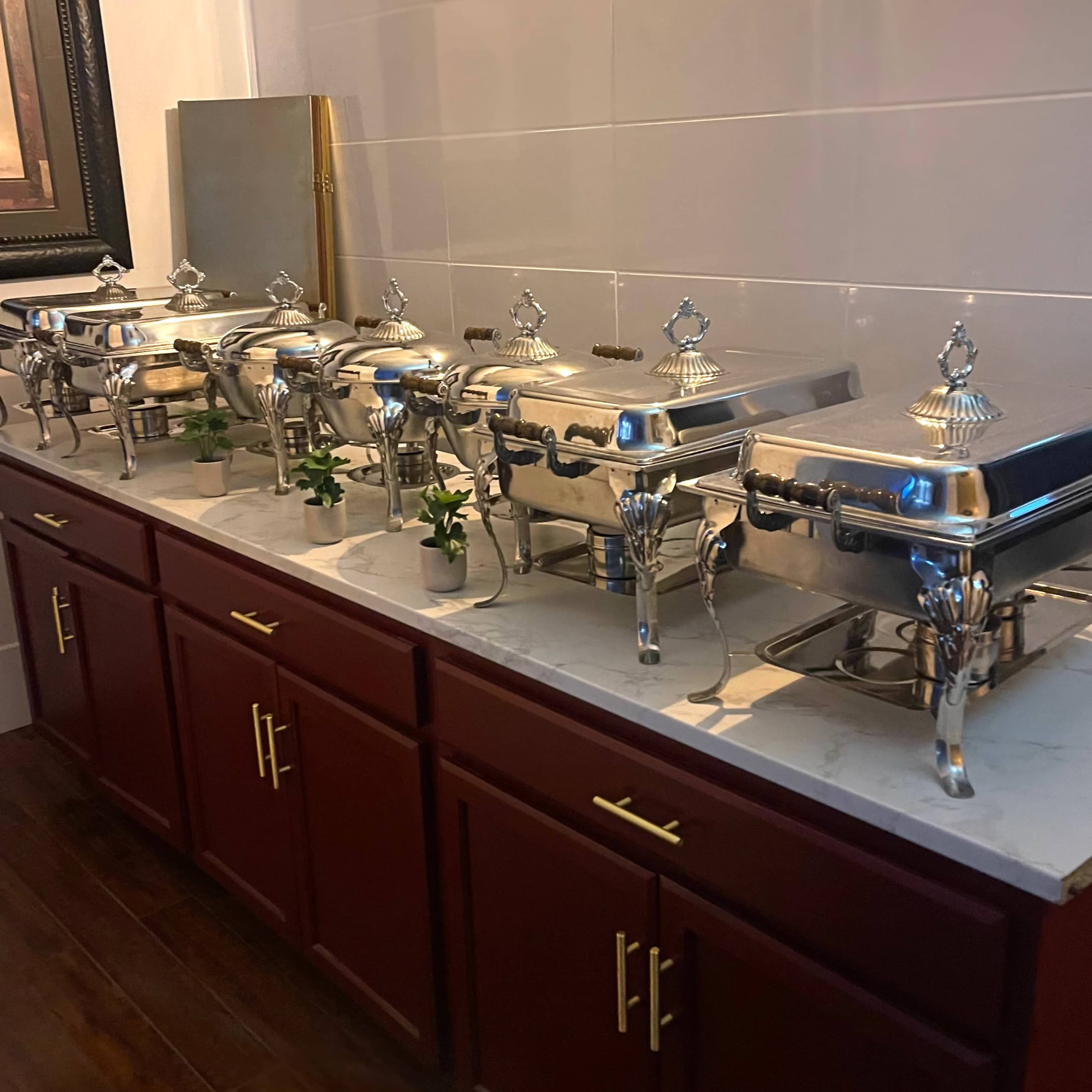 A row of silver serving chafing dishes is arranged on a wooden cabinet against a tiled wall.