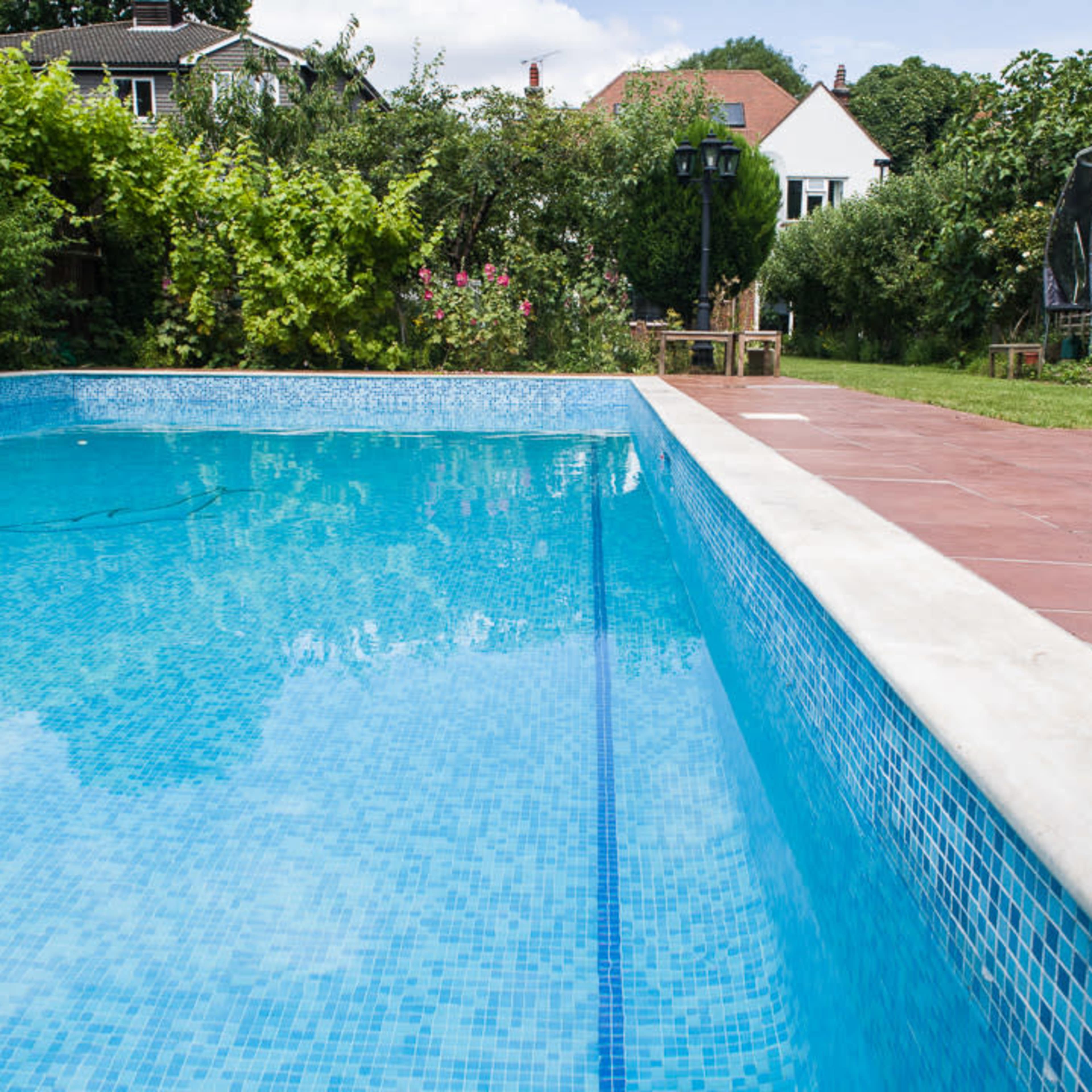 A clear swimming pool surrounded by a landscaped garden with a trampoline and seating area.