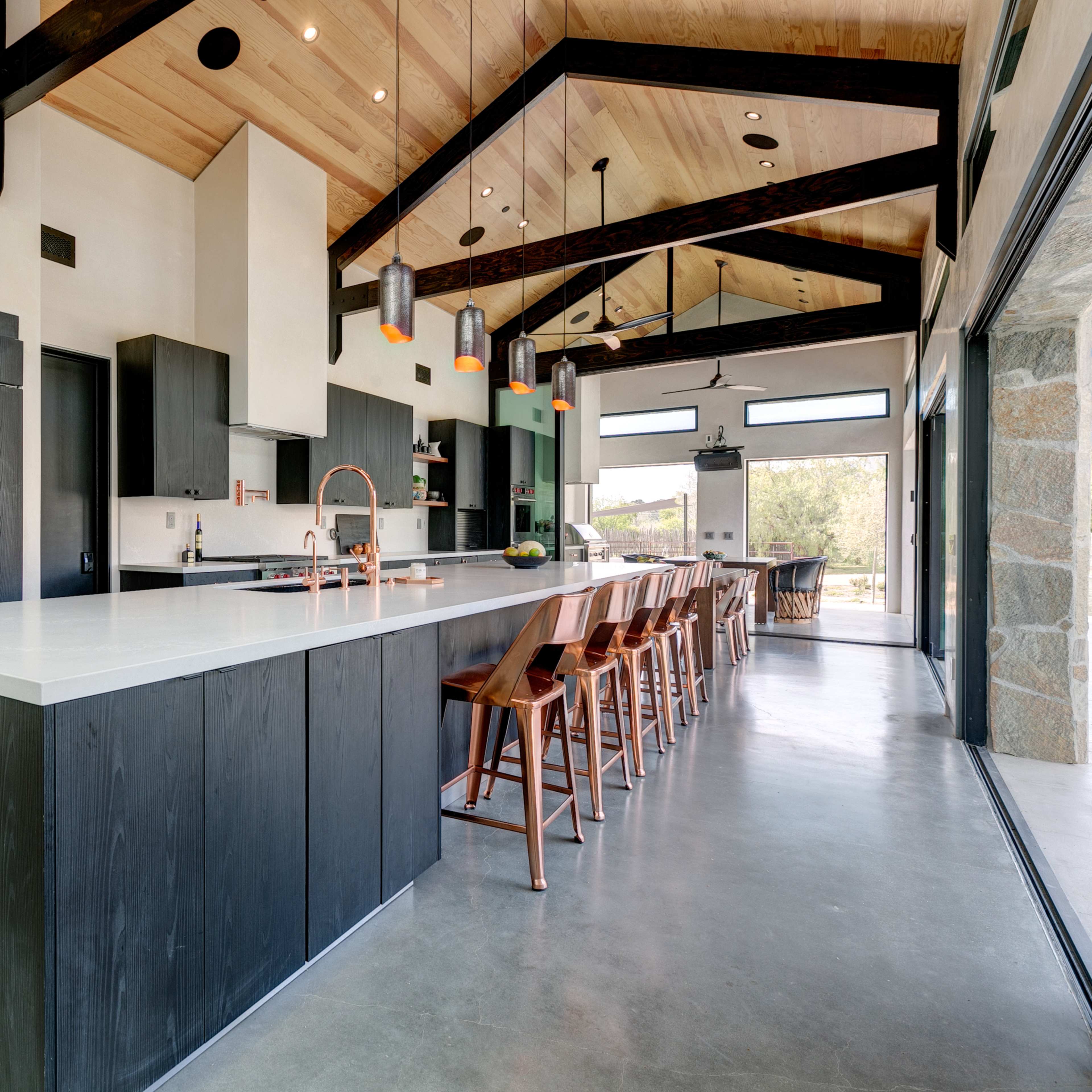 A modern kitchen features a large central island with a white countertop, black cabinetry, and wooden bar stools under a vaulted ceiling with exposed beams.
