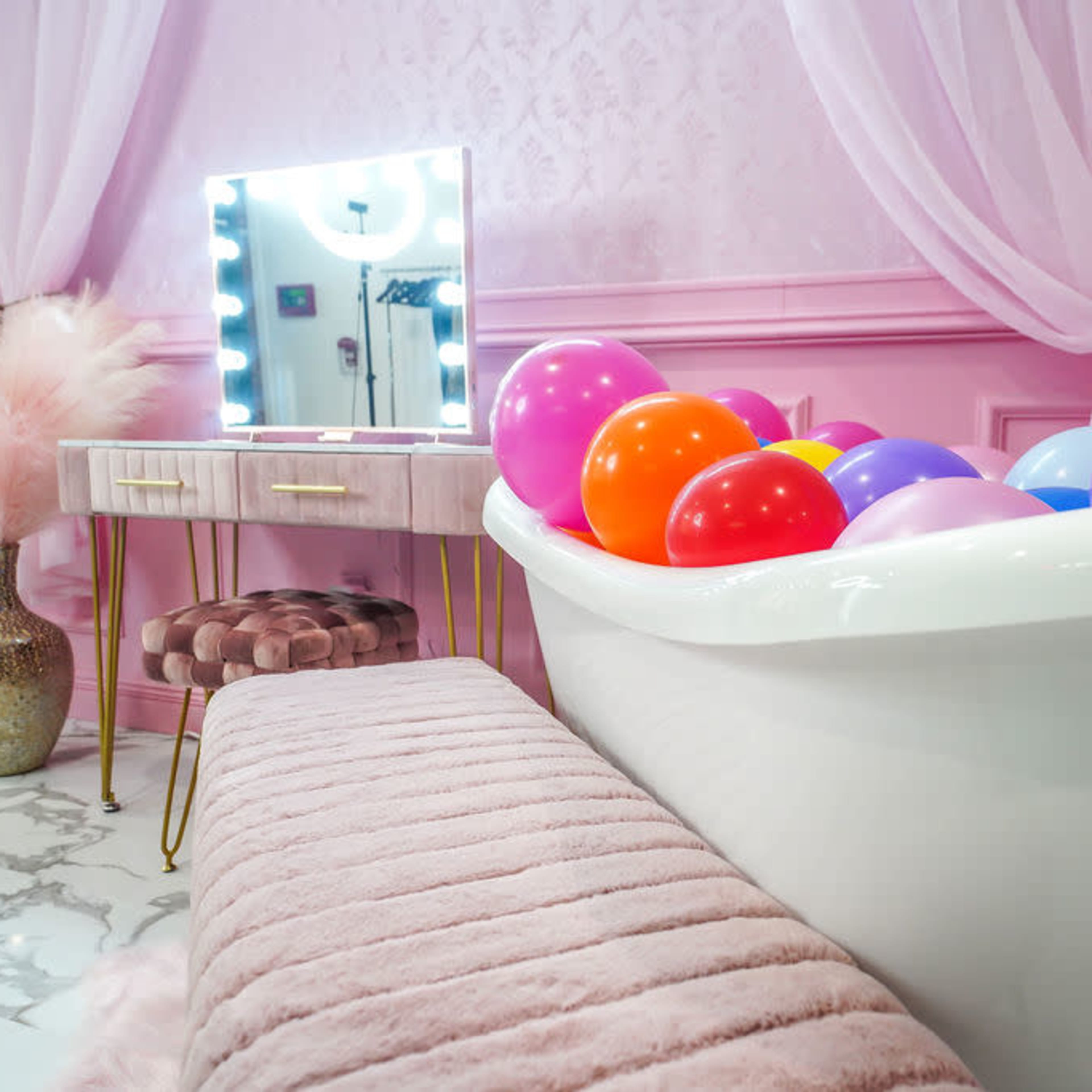 A white bathtub filled with colorful balloons is positioned next to a vanity with a mirror and a pink, plush bench in a elegantly decorated room.