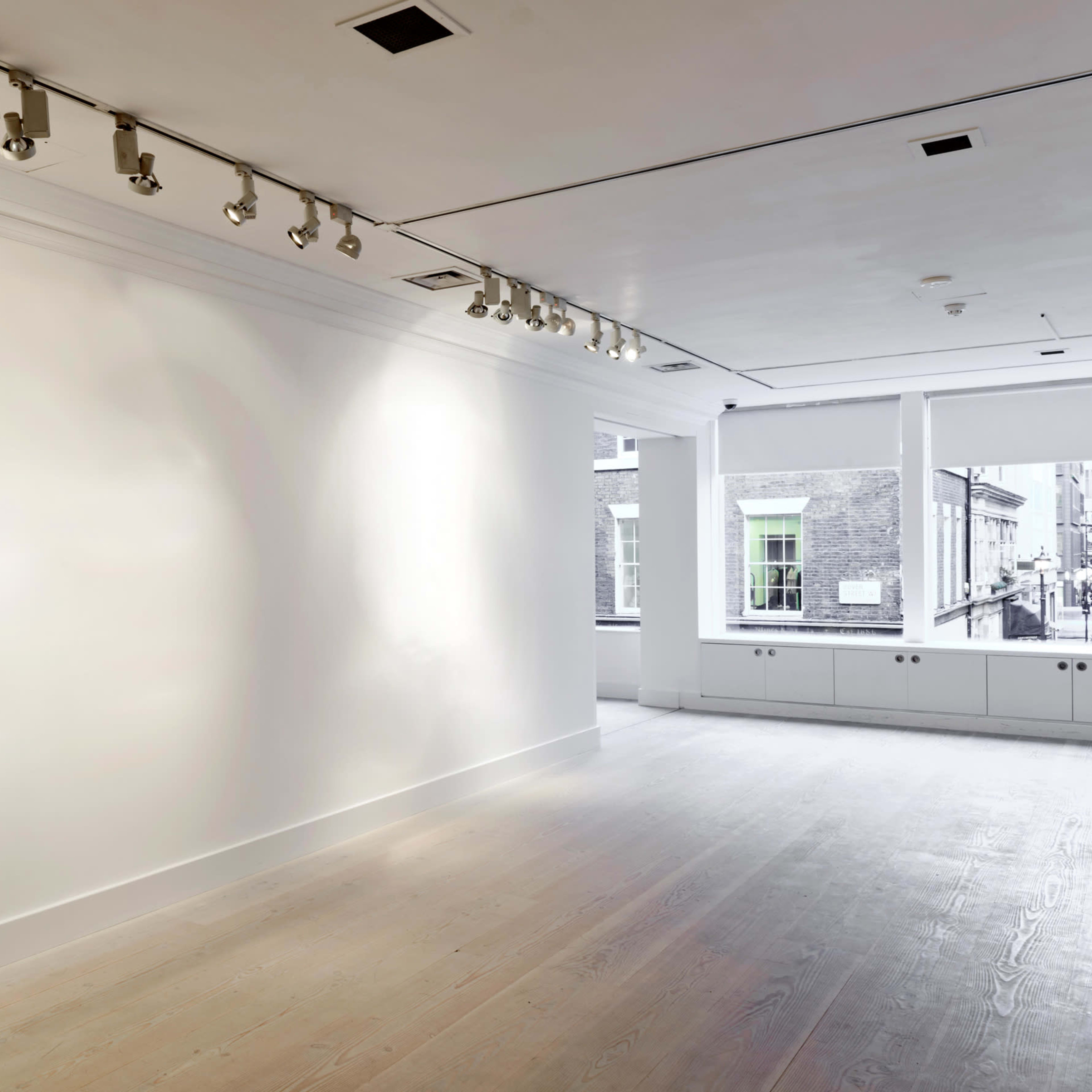 A large, empty room with white walls and wooden flooring, featuring track lighting on the ceiling and windows overlooking a street.