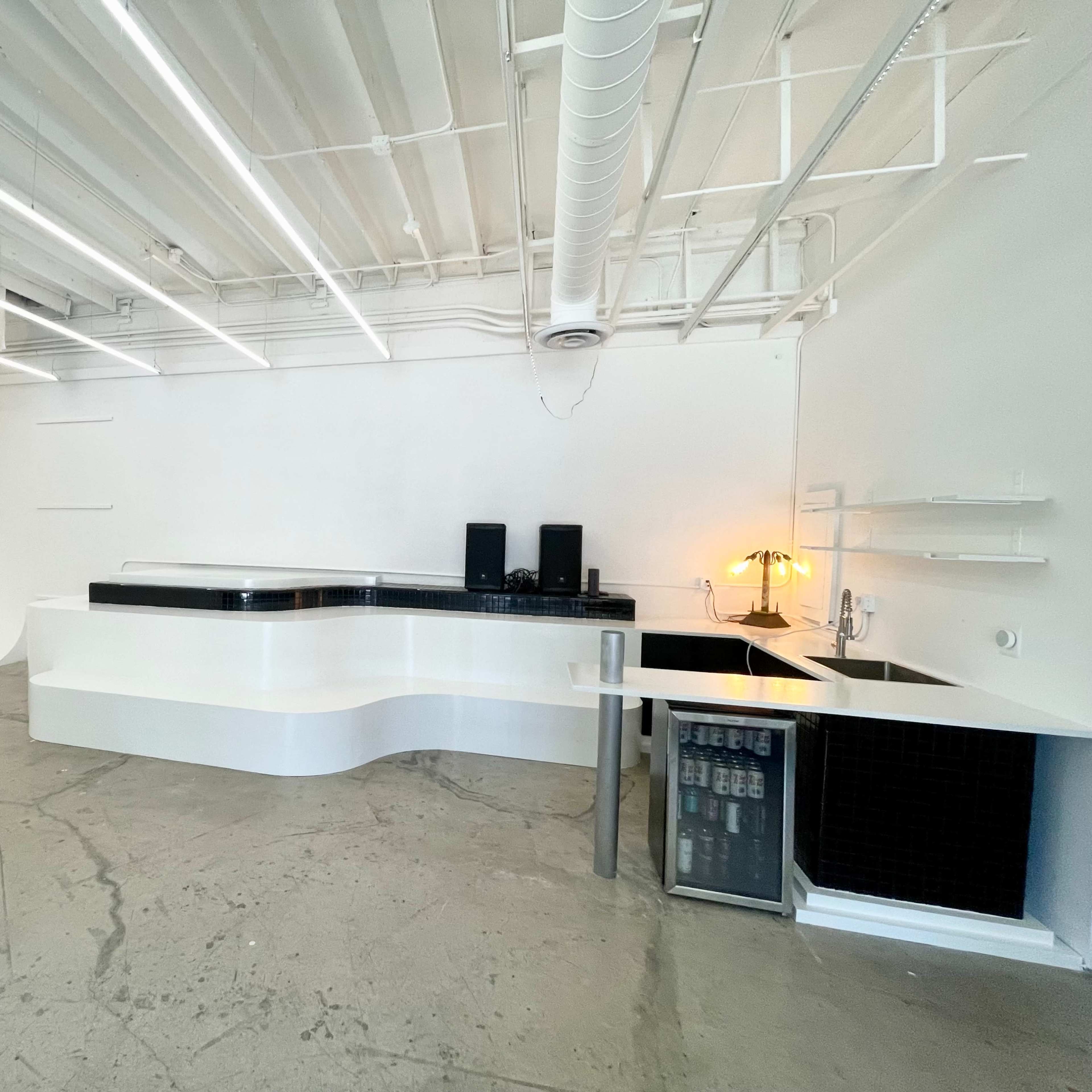 A modern, minimalistic interior space featuring a curved white reception counter, two black speakers, and a small beverage refrigerator against a plain wall.
