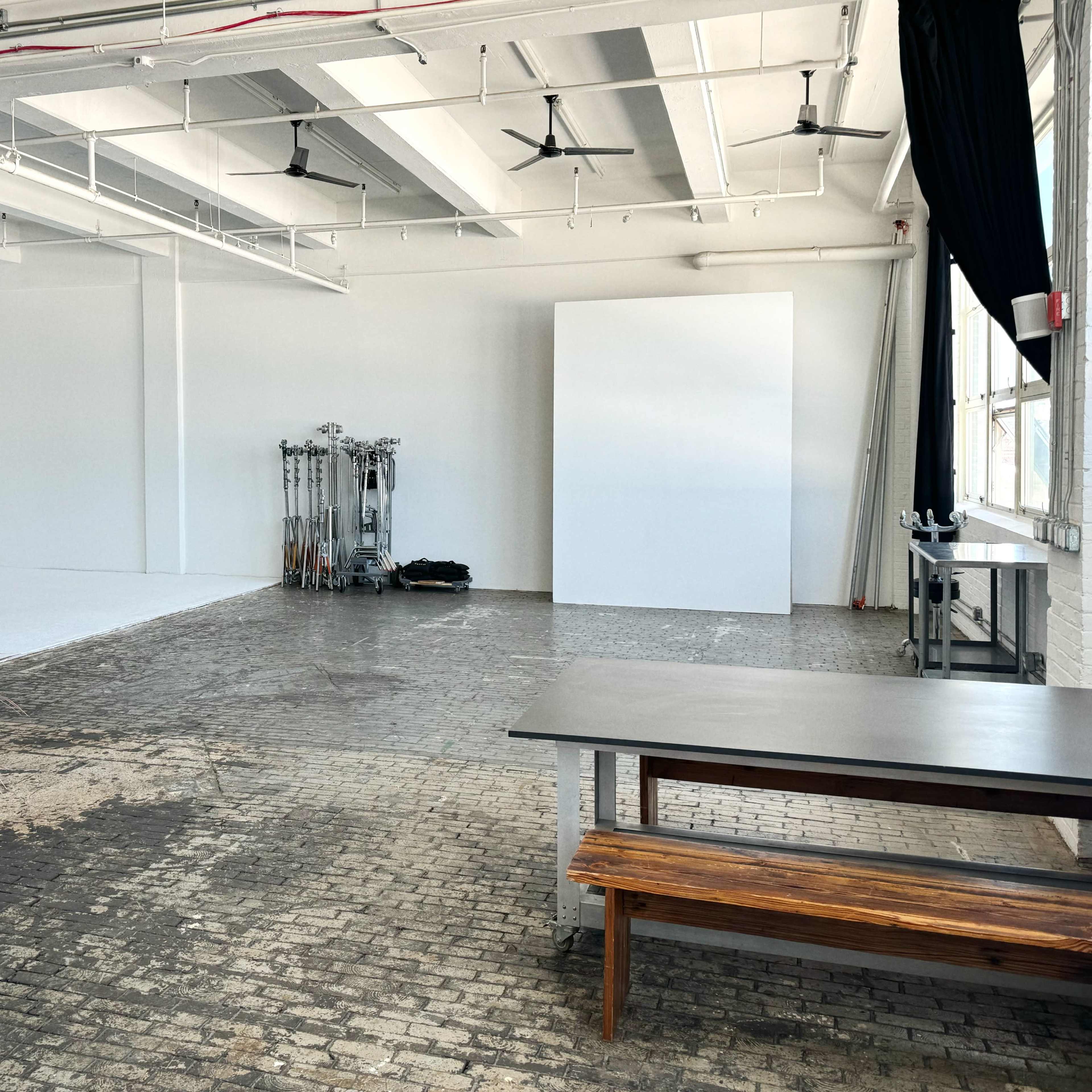 A spacious, empty studio with brick flooring, large windows, and minimal furnishings, including a potted plant and two wooden tables.