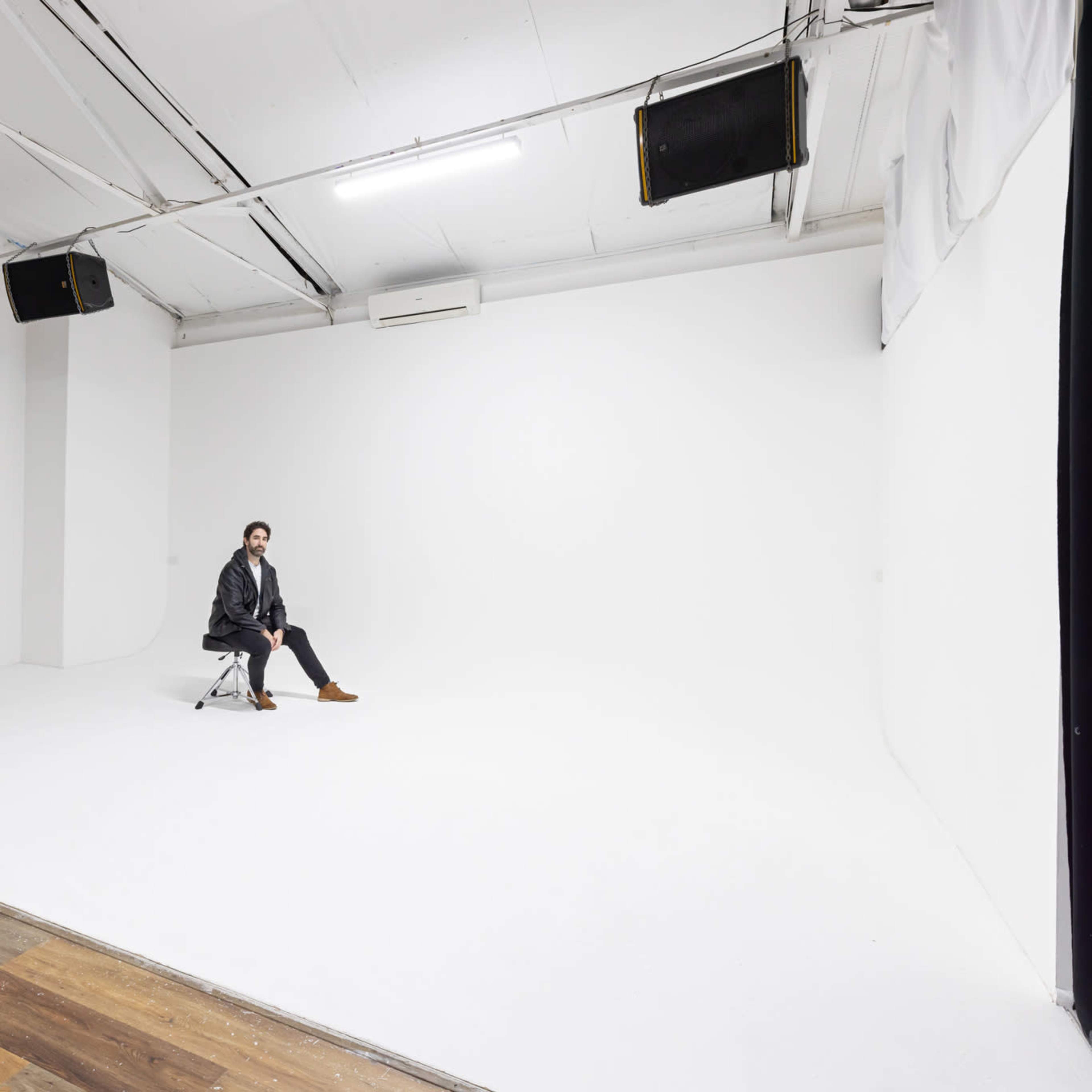 A person sits on a chair in an empty, well-lit studio with white walls and a wooden floor.
