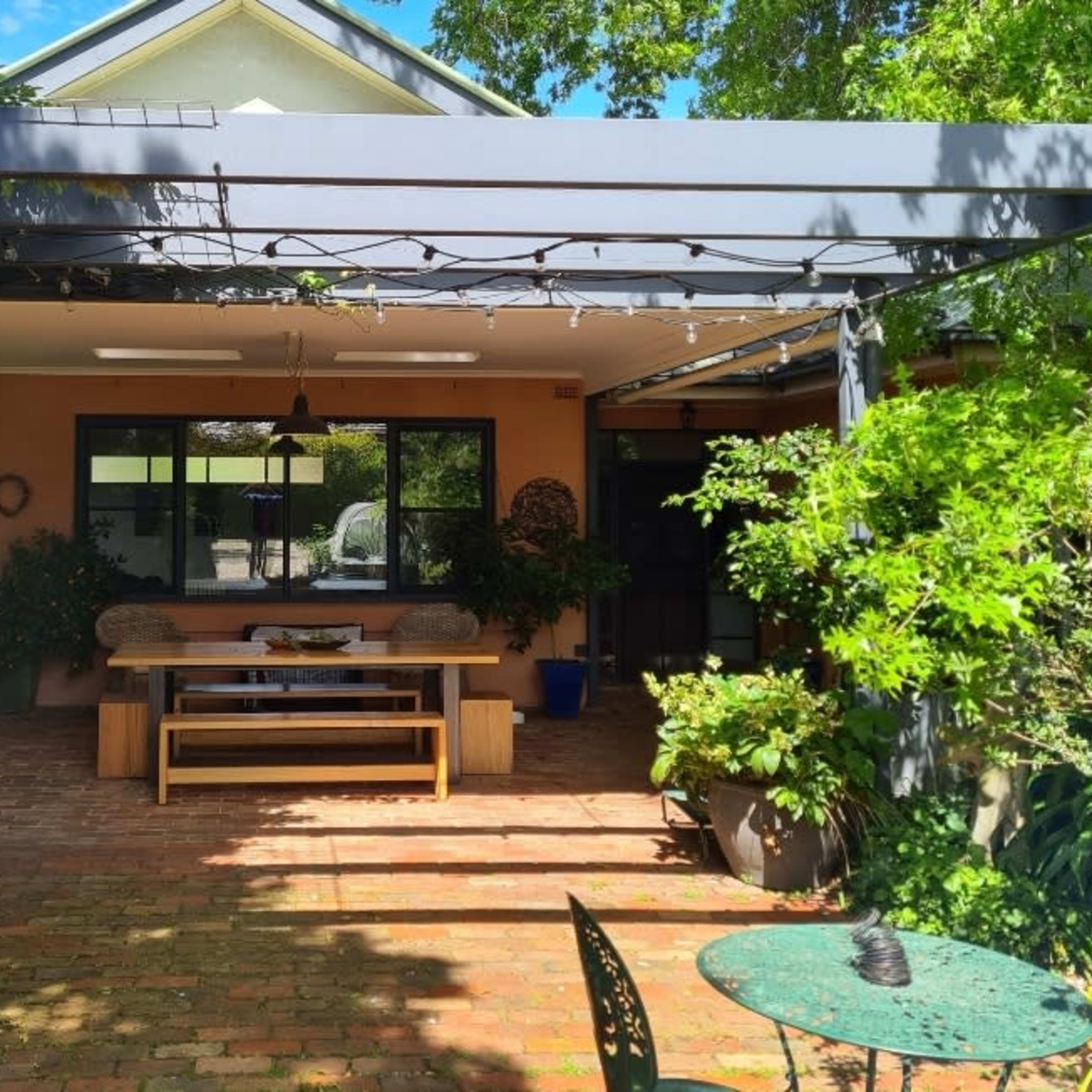 A covered outdoor patio features a wooden table and benches surrounded by greenery and decorative plants.