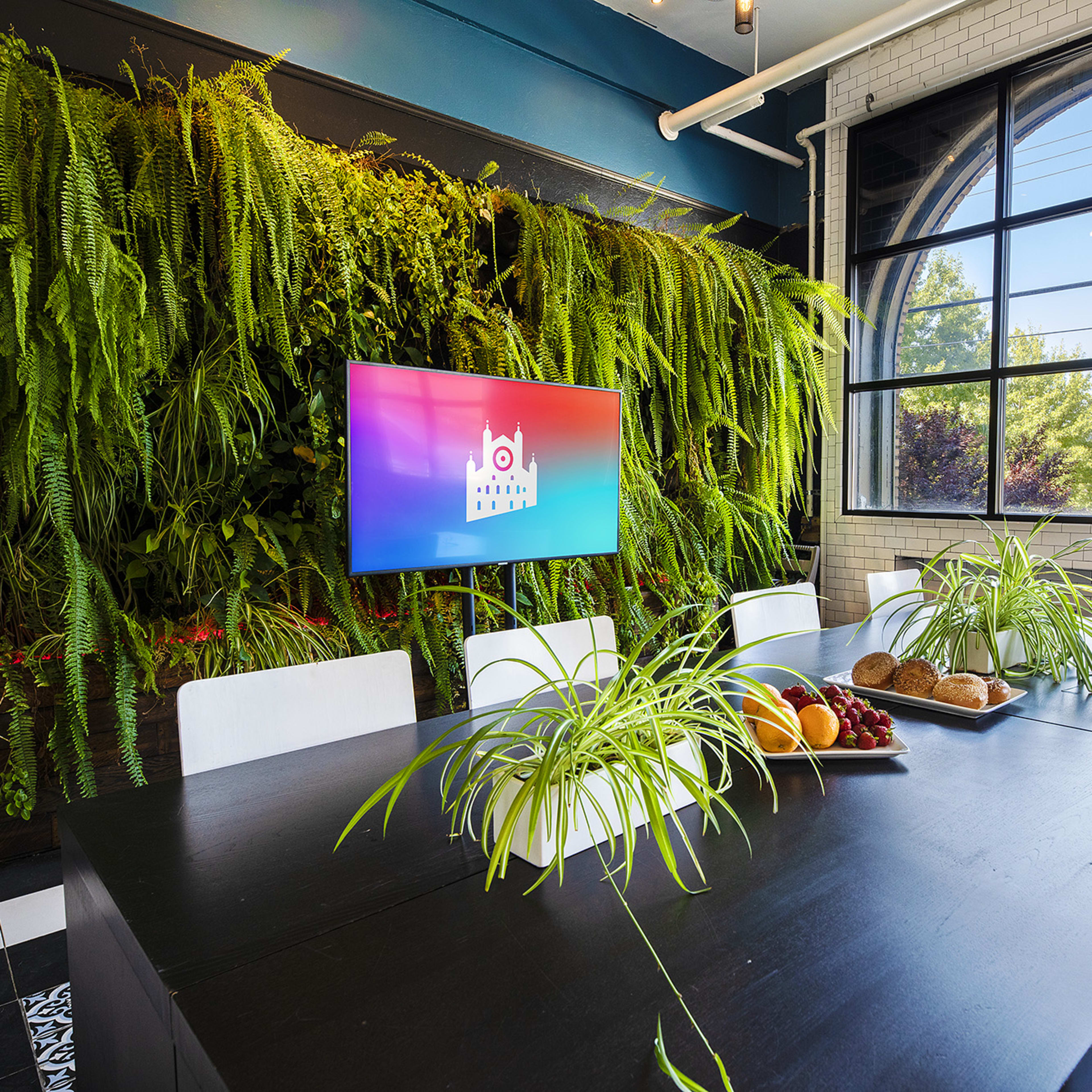 A large meeting room features a wall of greenery, a digital screen, and a dining table set with fruits and baked goods.