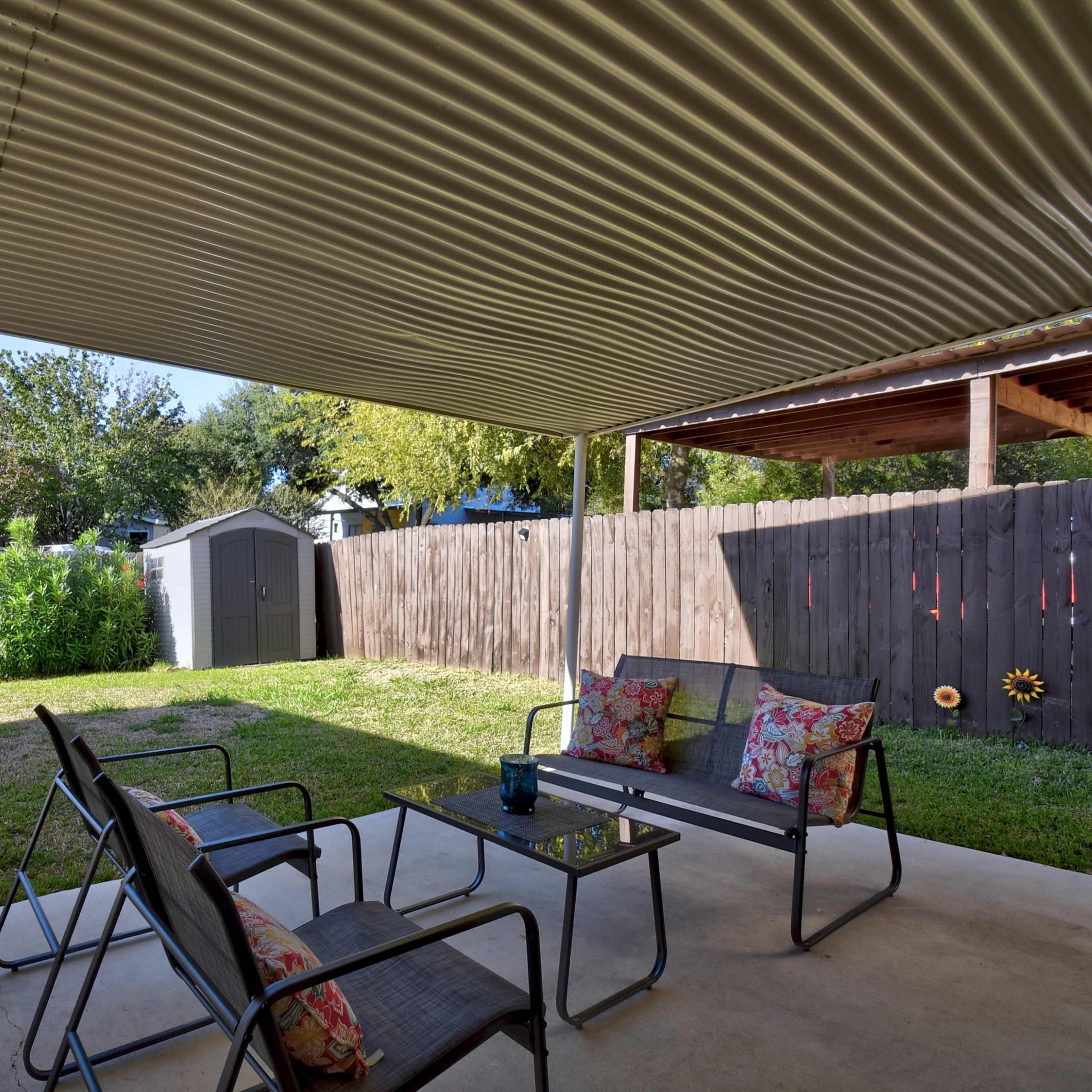 A covered patio features metal chairs with cushions facing a grassy backyard and a storage shed.