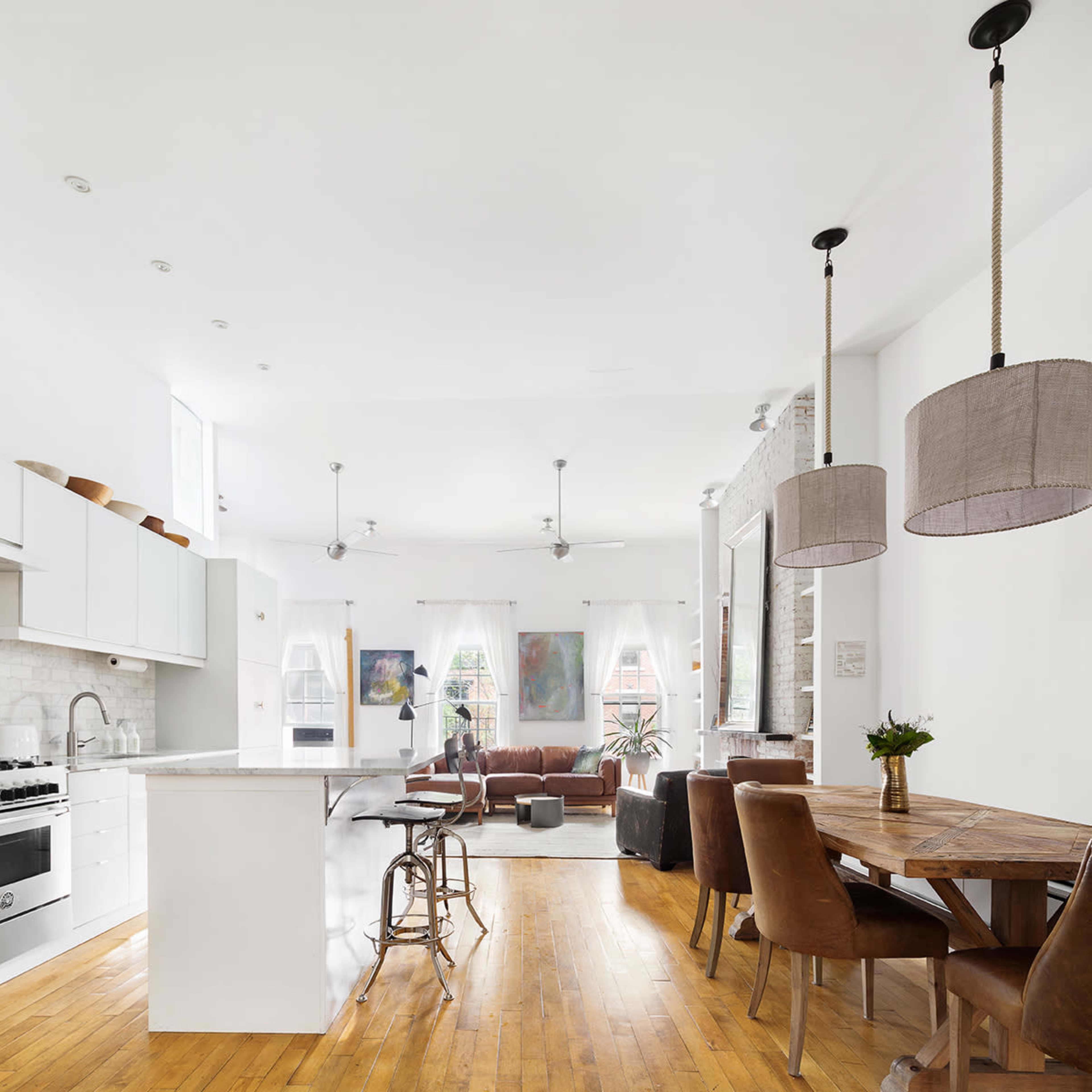 A spacious kitchen and living area featuring modern appliances, wooden floors, a dining table, and large windows with natural light.
