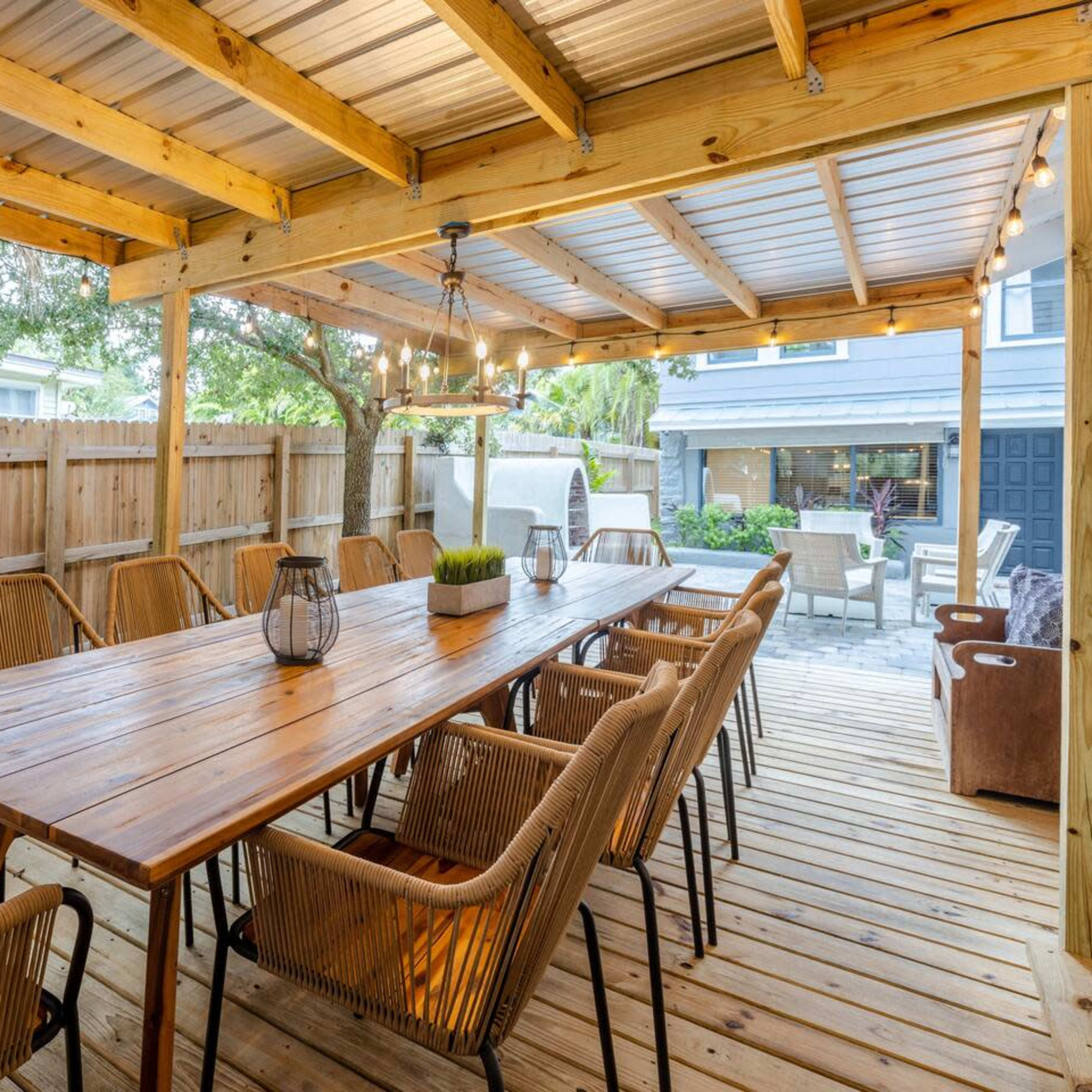 A wooden patio with a long table surrounded by chairs, under a covered structure with string lights.