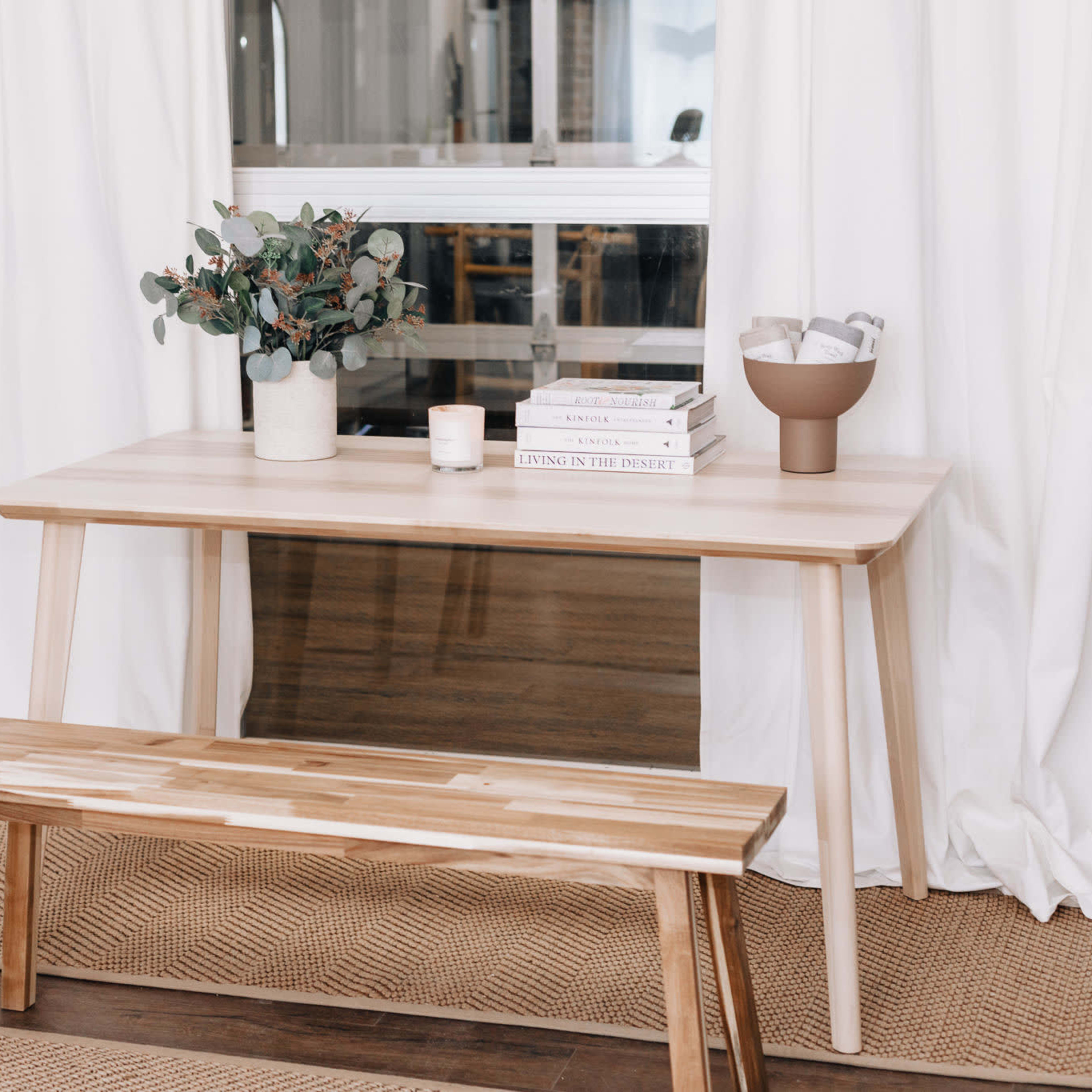 A modern wooden table with a bench sits on a woven rug, featuring a vase of greenery and a stack of books beside a candle.