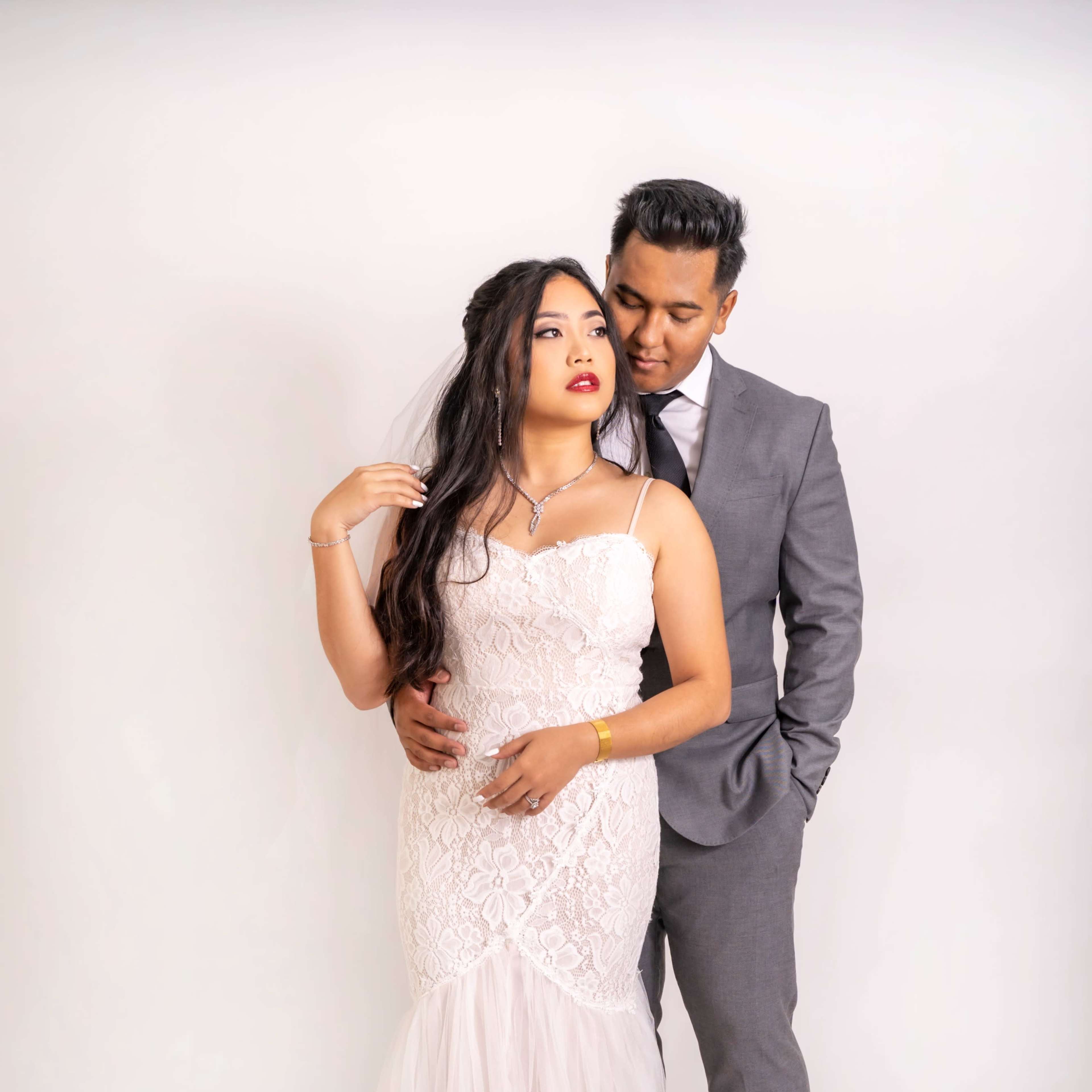 A bride in a lace wedding dress stands next to a man in a gray suit, both posing together against a light background.