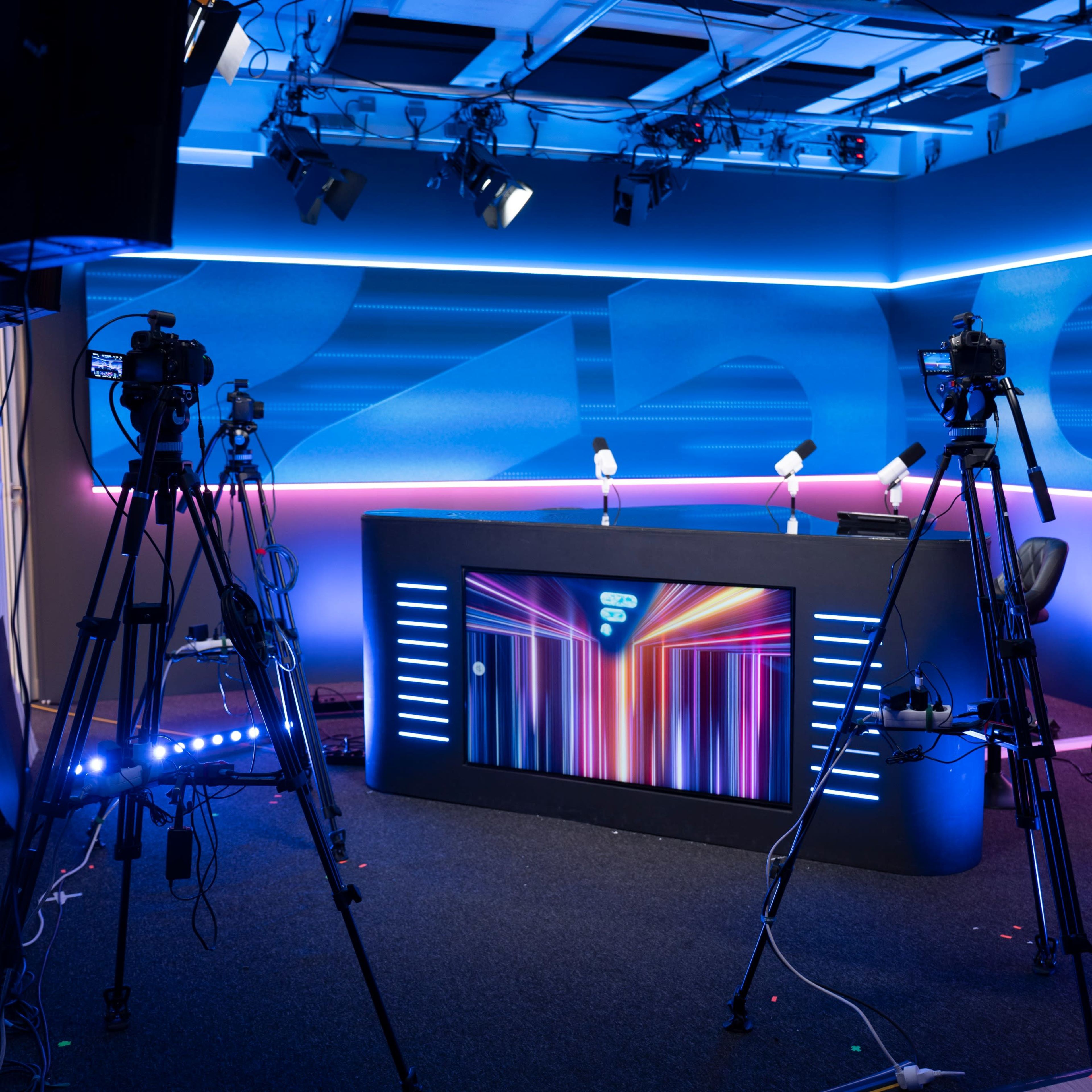 A brightly lit television studio features a central desk with colorful lighting and multiple cameras positioned around it.