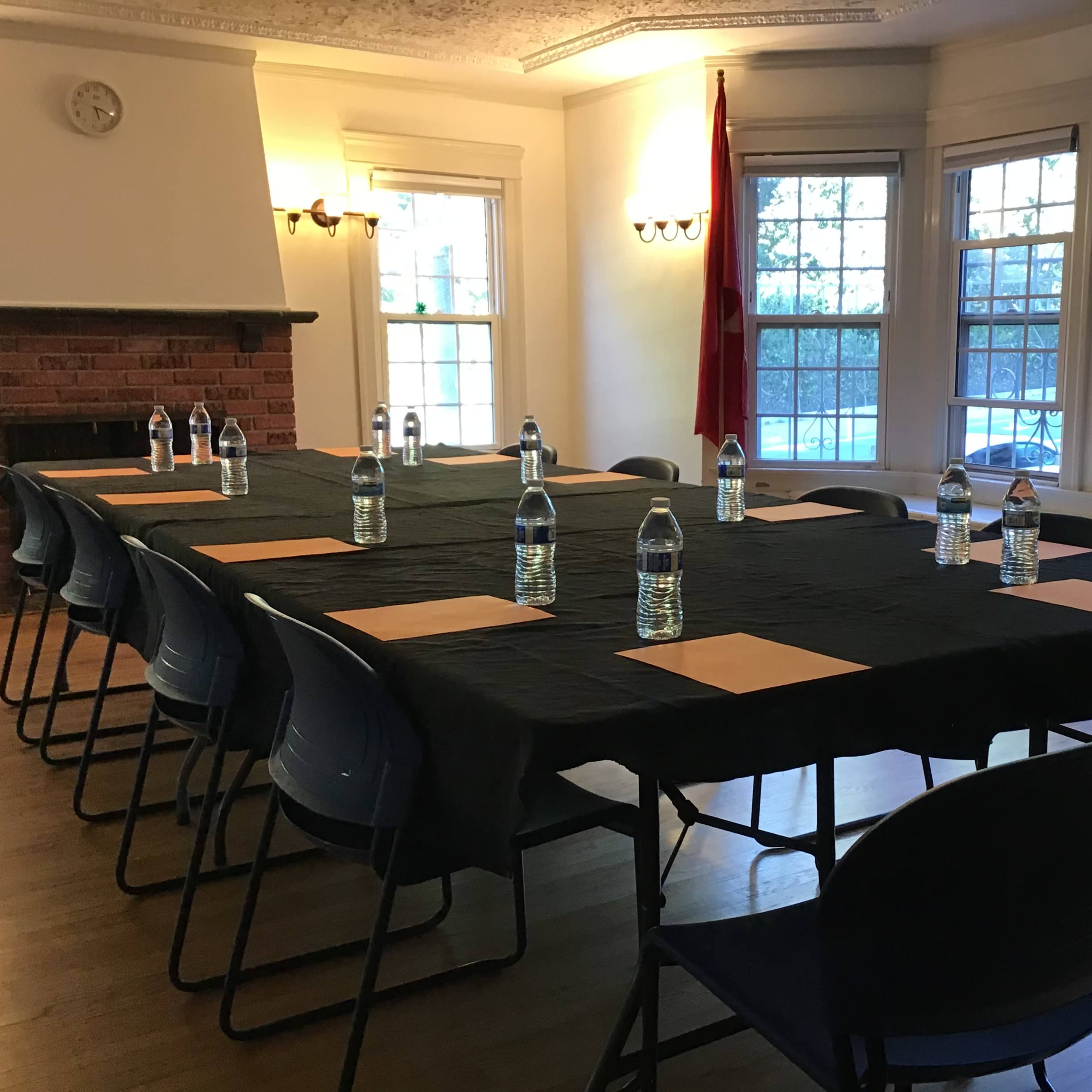 A long table is set with water bottles and paper placemats in a well-lit room featuring large windows and a brick fireplace.