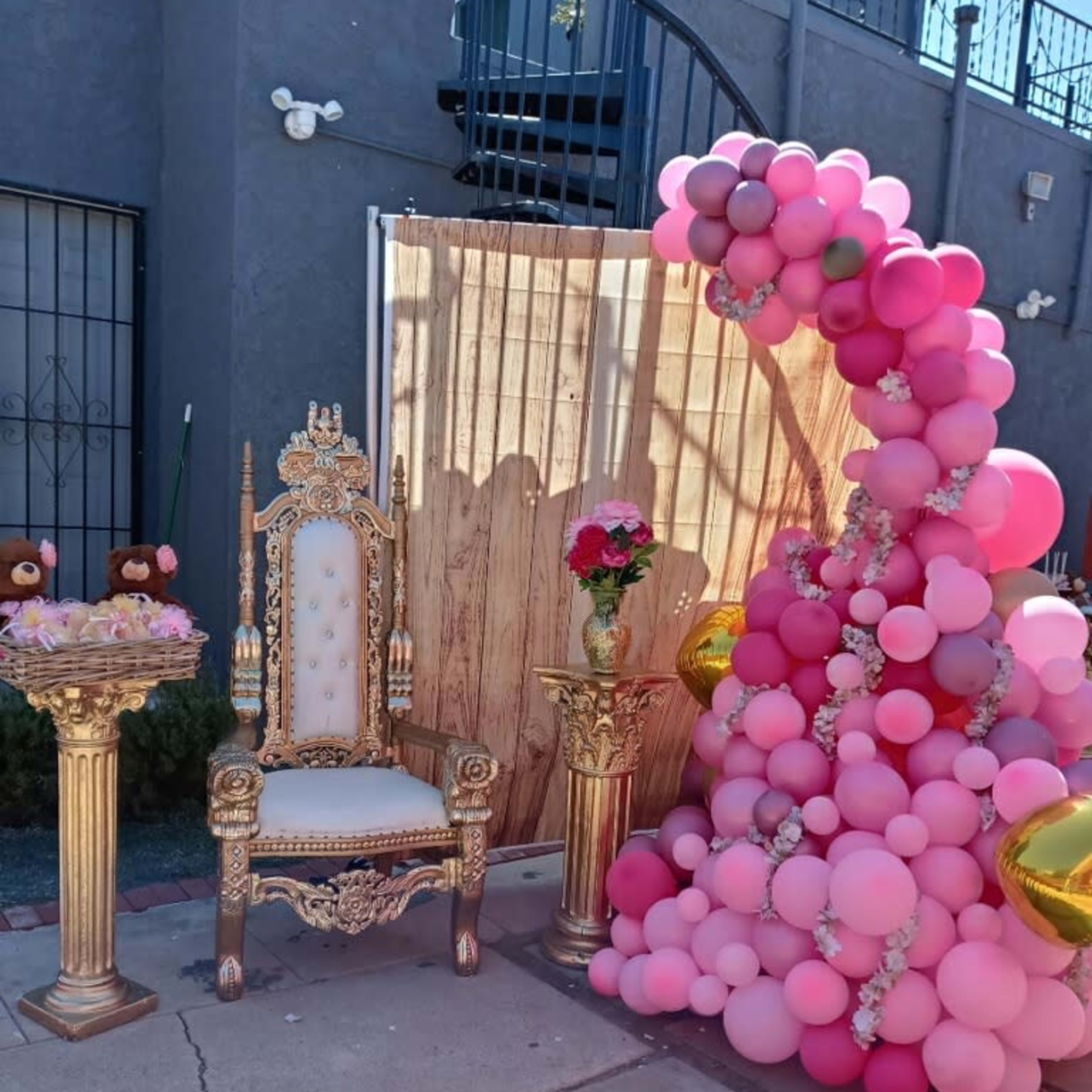 The image shows a decorated party corner featuring a golden throne, two pedestal tables with floral arrangements, and a large arch of pink balloons.