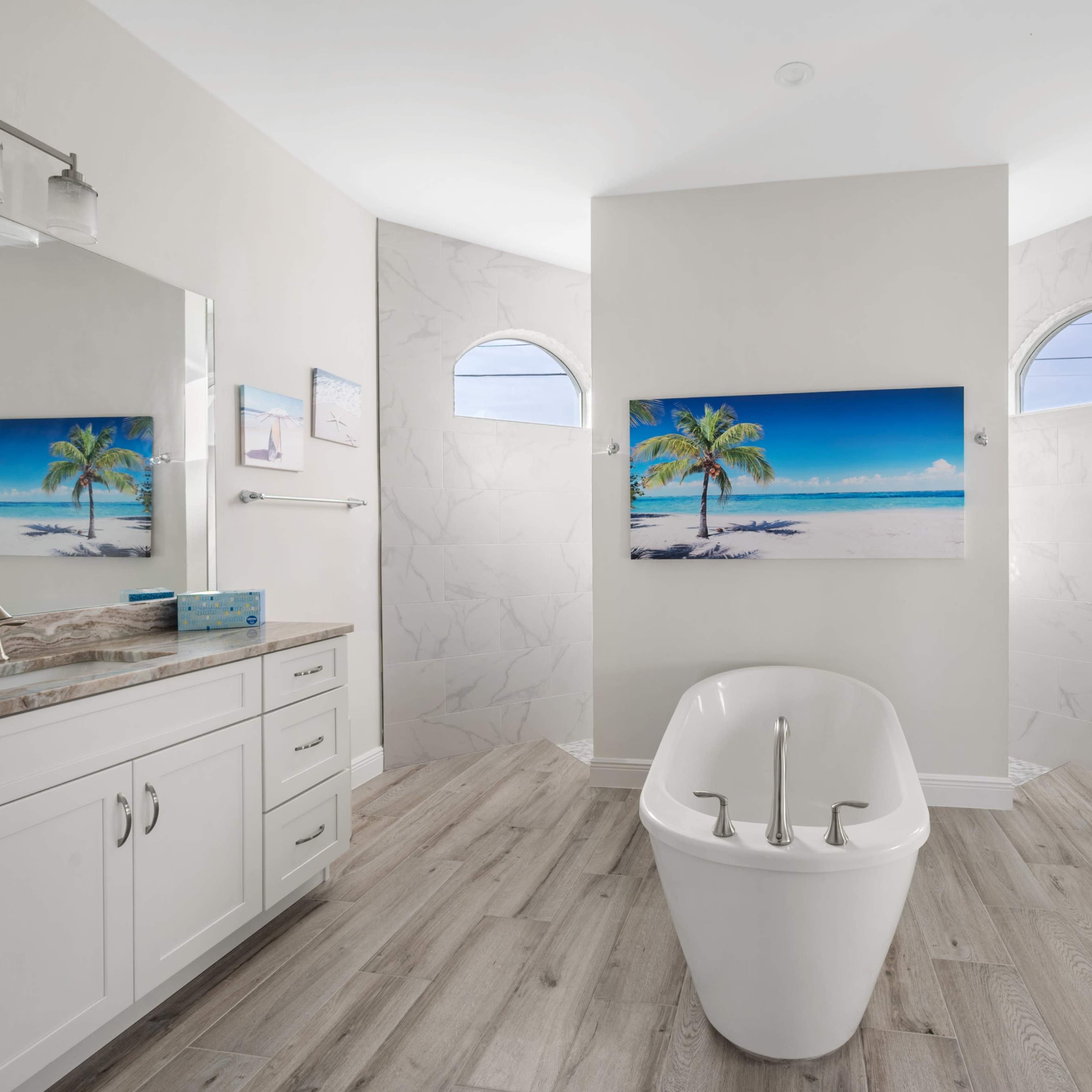 A modern bathroom features a freestanding bathtub, large mirrors, and a beach-themed artwork on the wall, with light-colored flooring and walls.