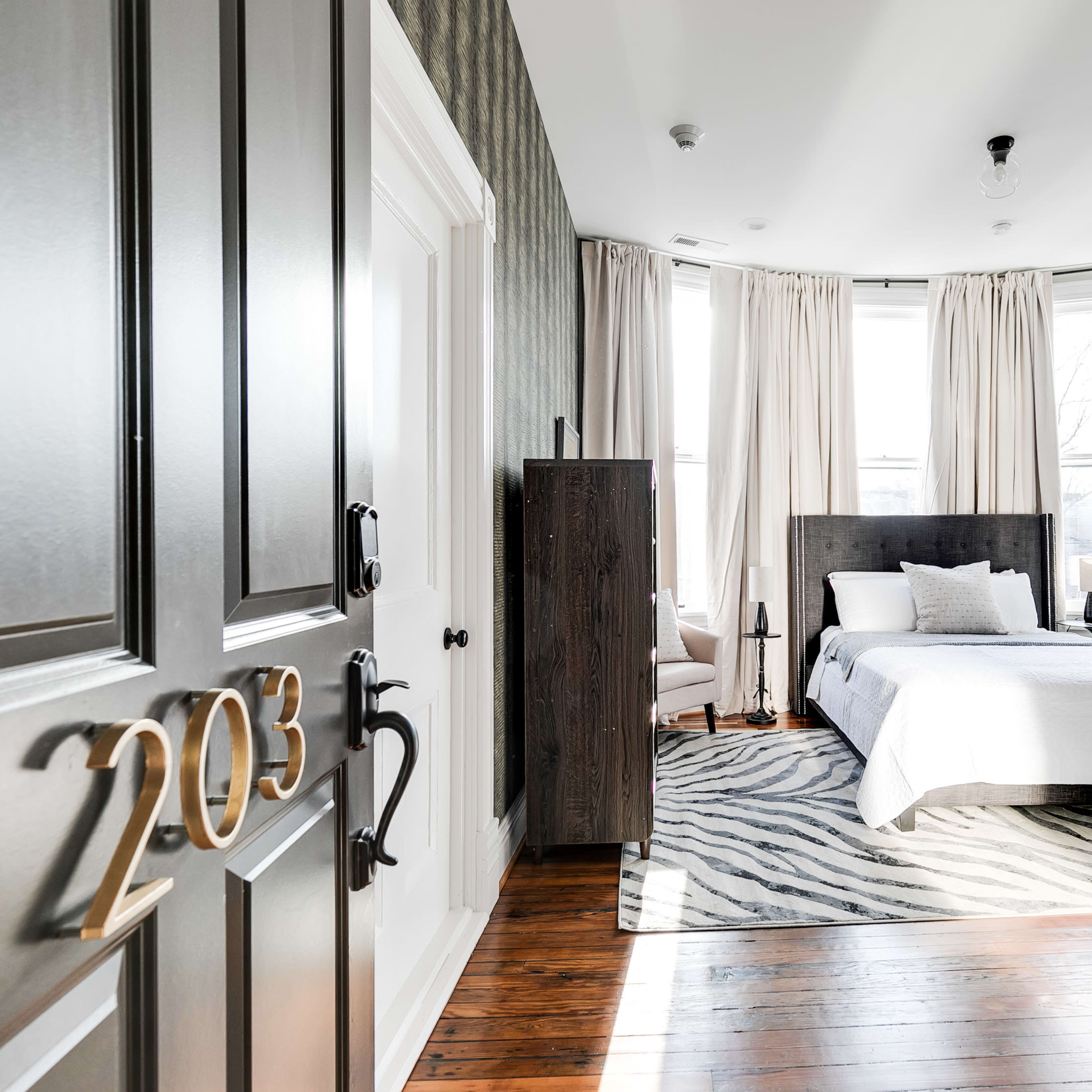 A modern hotel room with a bed, large windows, and a door marked "203" in a corridor.