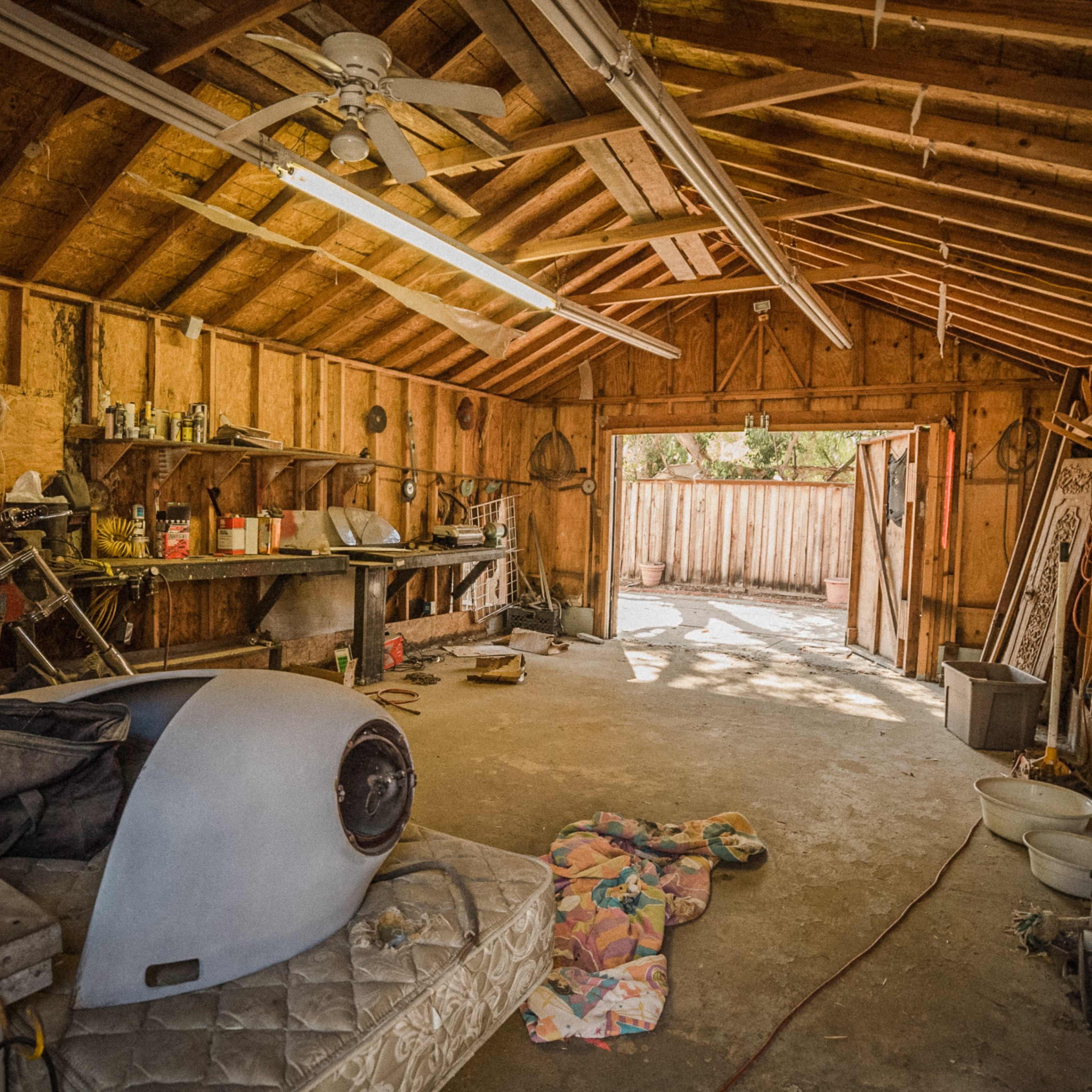 The interior of a wooden garage with unfinished walls, featuring a workbench, tools, and an open door leading to an outdoor area.