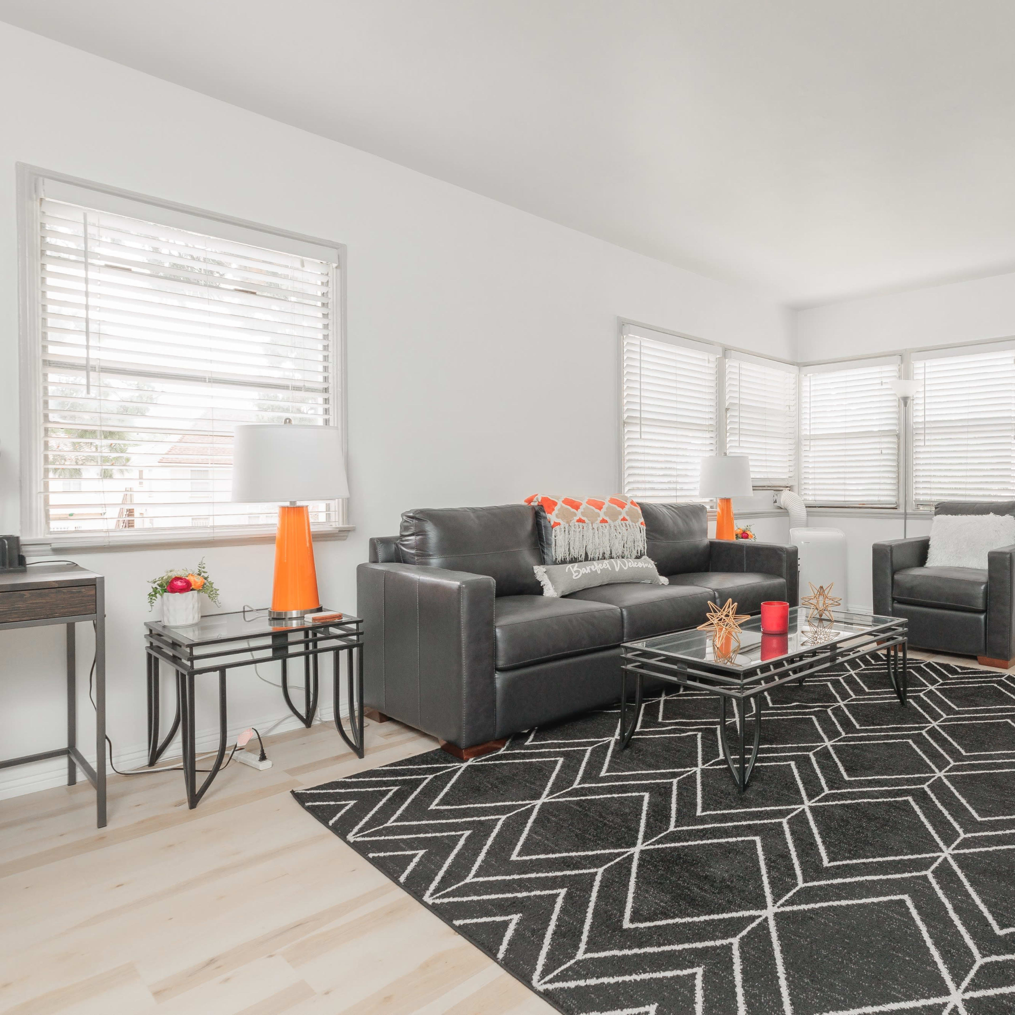 A modern living room features a black and white geometric rug, a glass coffee table, and a gray seating arrangement with bright accents.