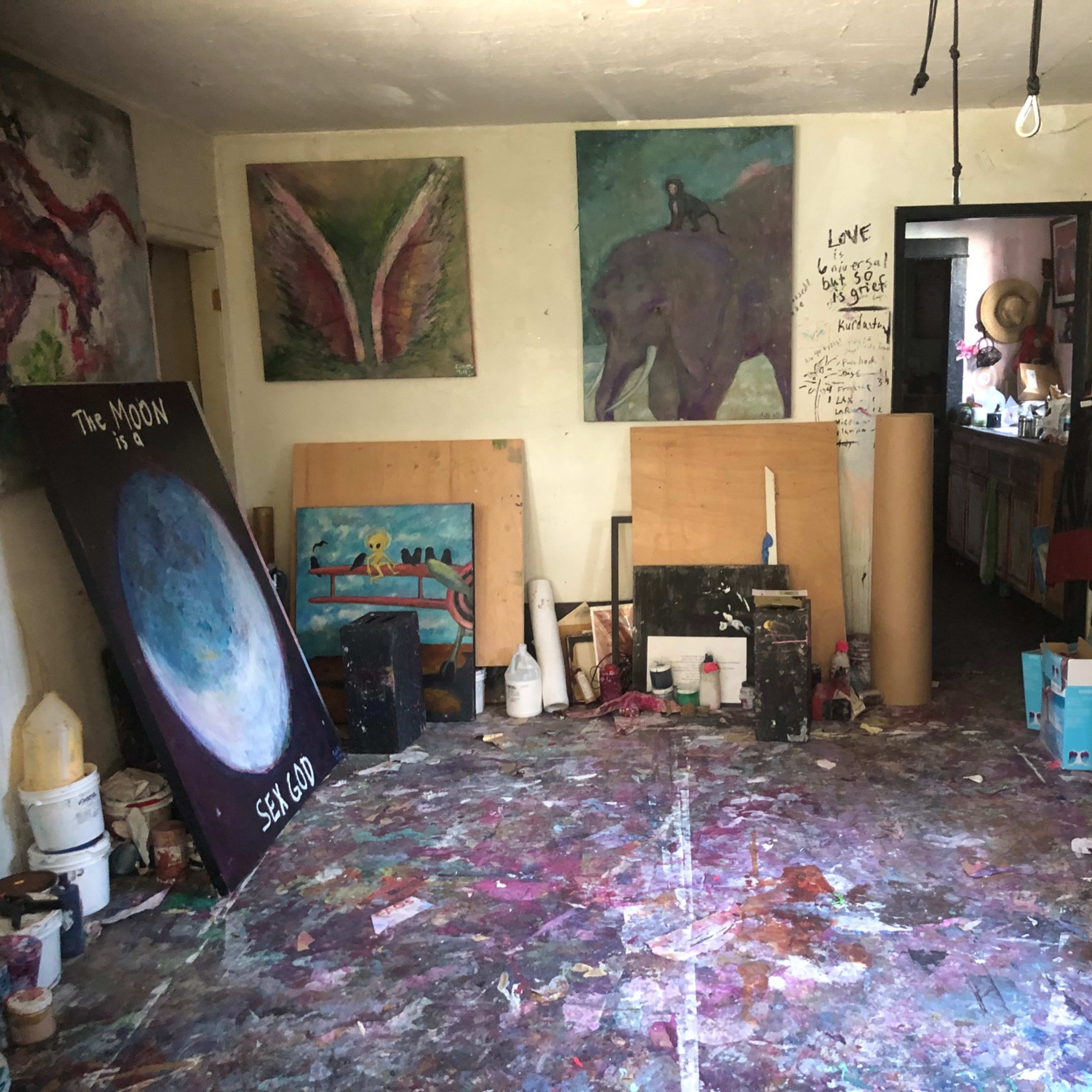A cluttered art studio with colorful paintings on the walls, scattered art supplies, and a messy floor covered in paint splatters.