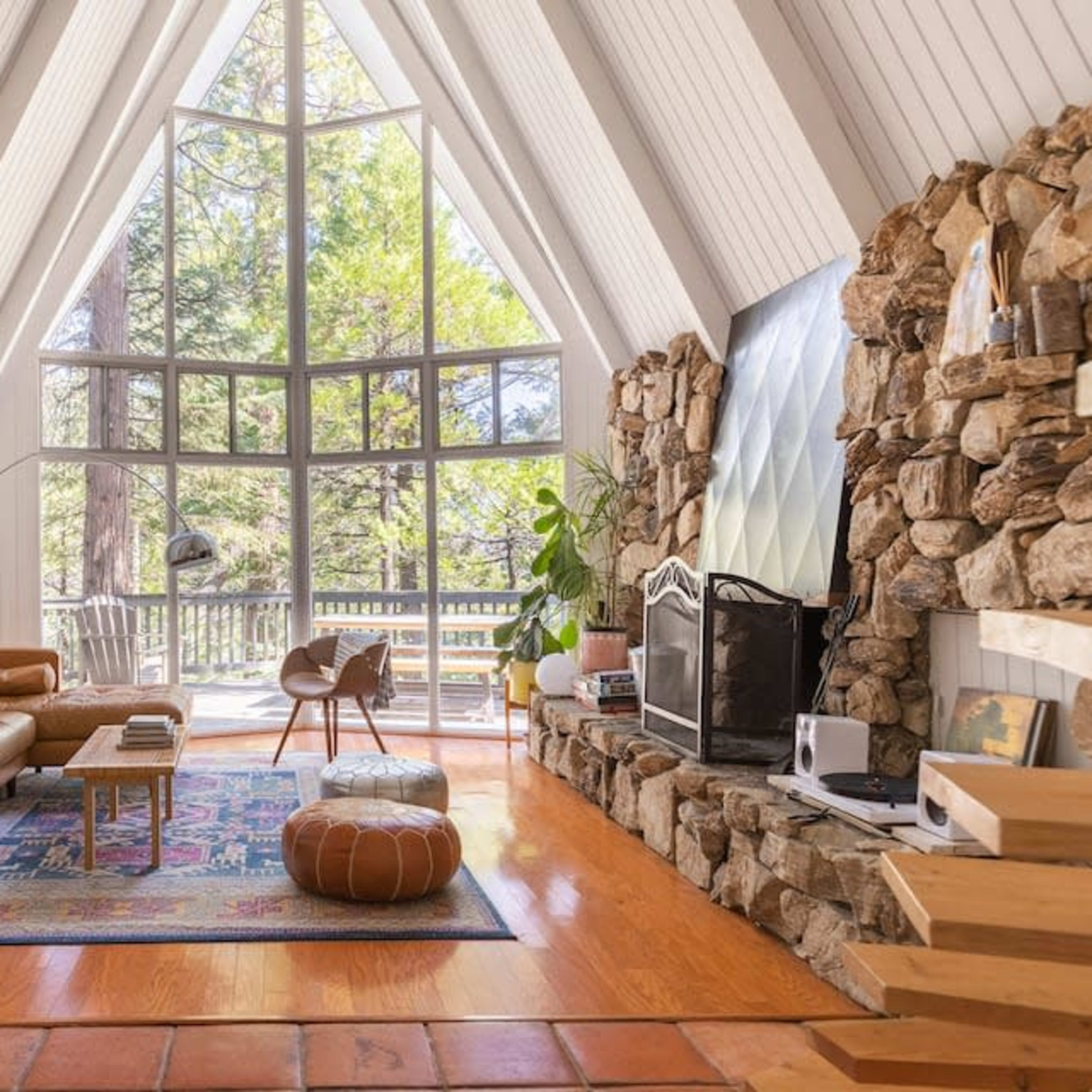 A cozy living room in an A-frame cabin with a stone fireplace, large windows letting in natural light, and a mix of wooden and modern furnishings.