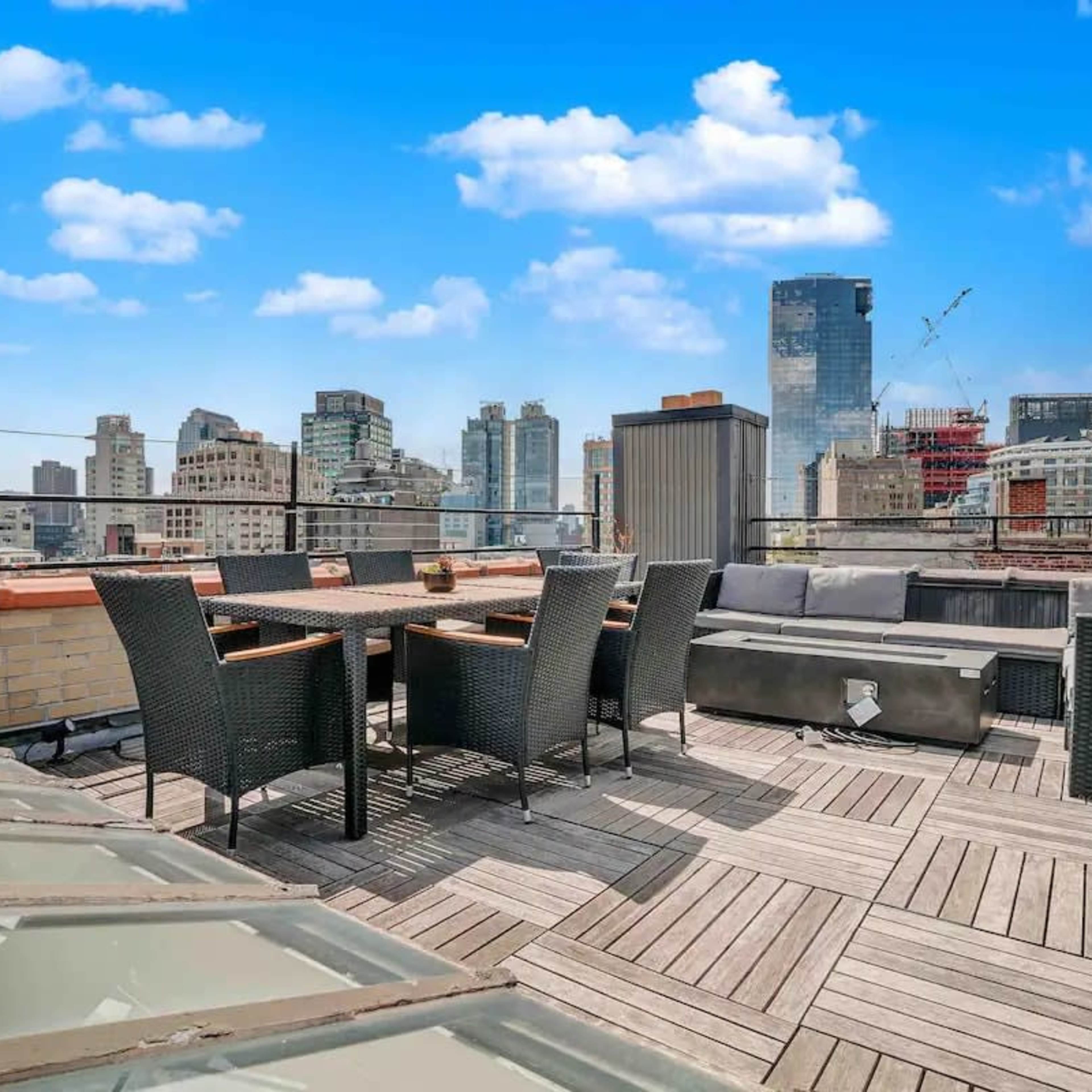 A rooftop terrace features a seating area with wicker furniture and a view of a city skyline under a blue sky.
