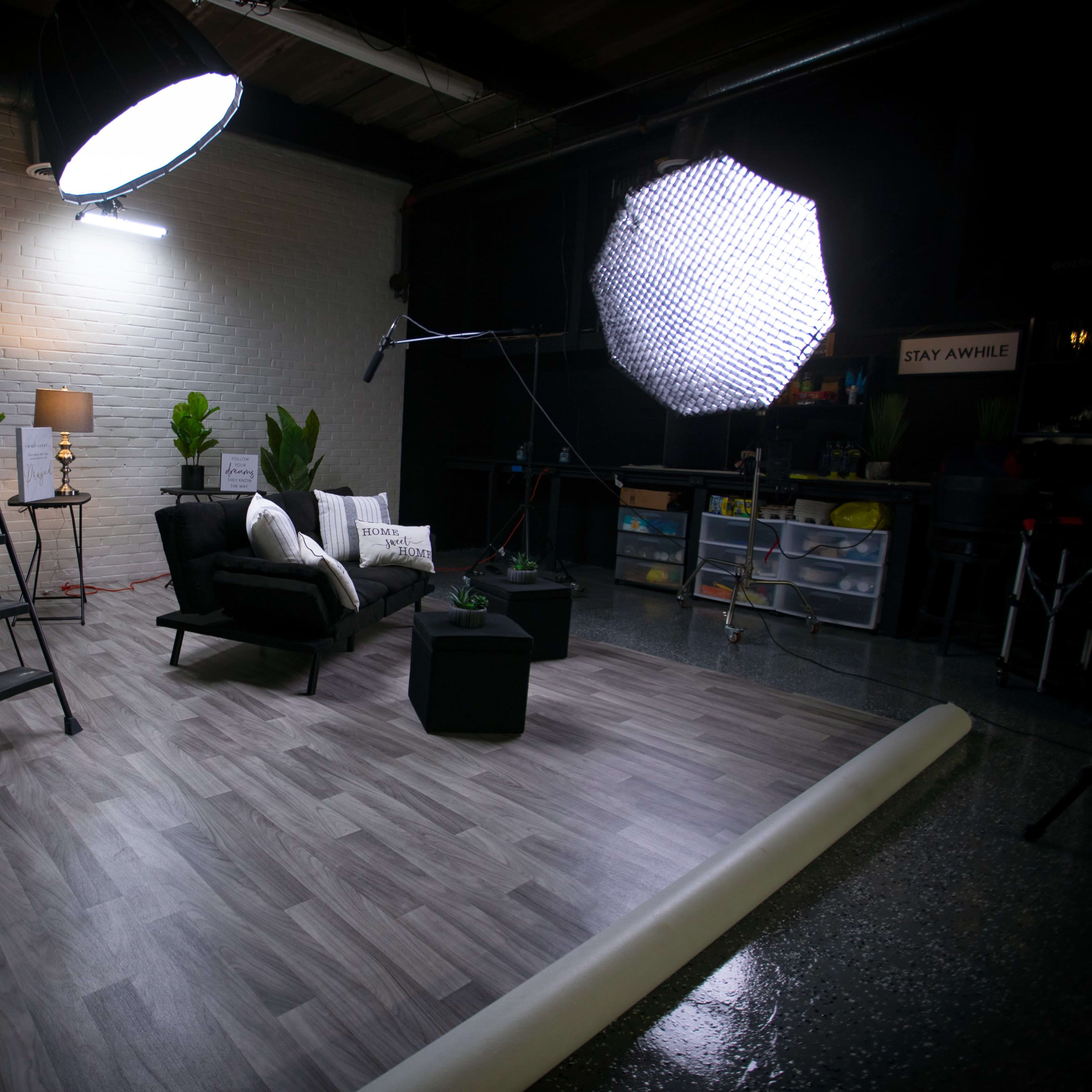 A staged indoor setting with two black chairs, a small table, plants, and lighting equipment on a wooden floor.