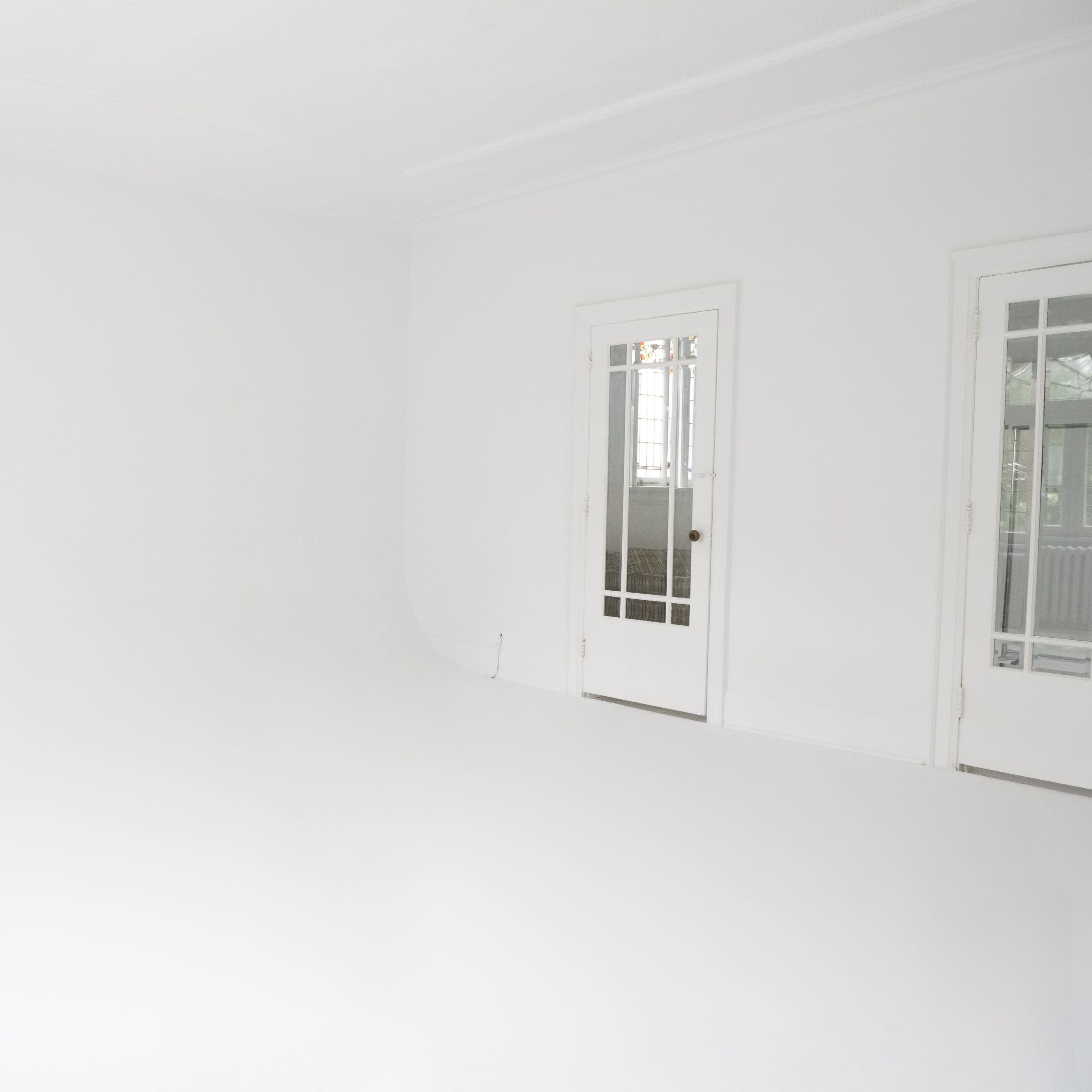 A spacious, empty room with white walls and a white floor, featuring large windows and a door leading to another room.