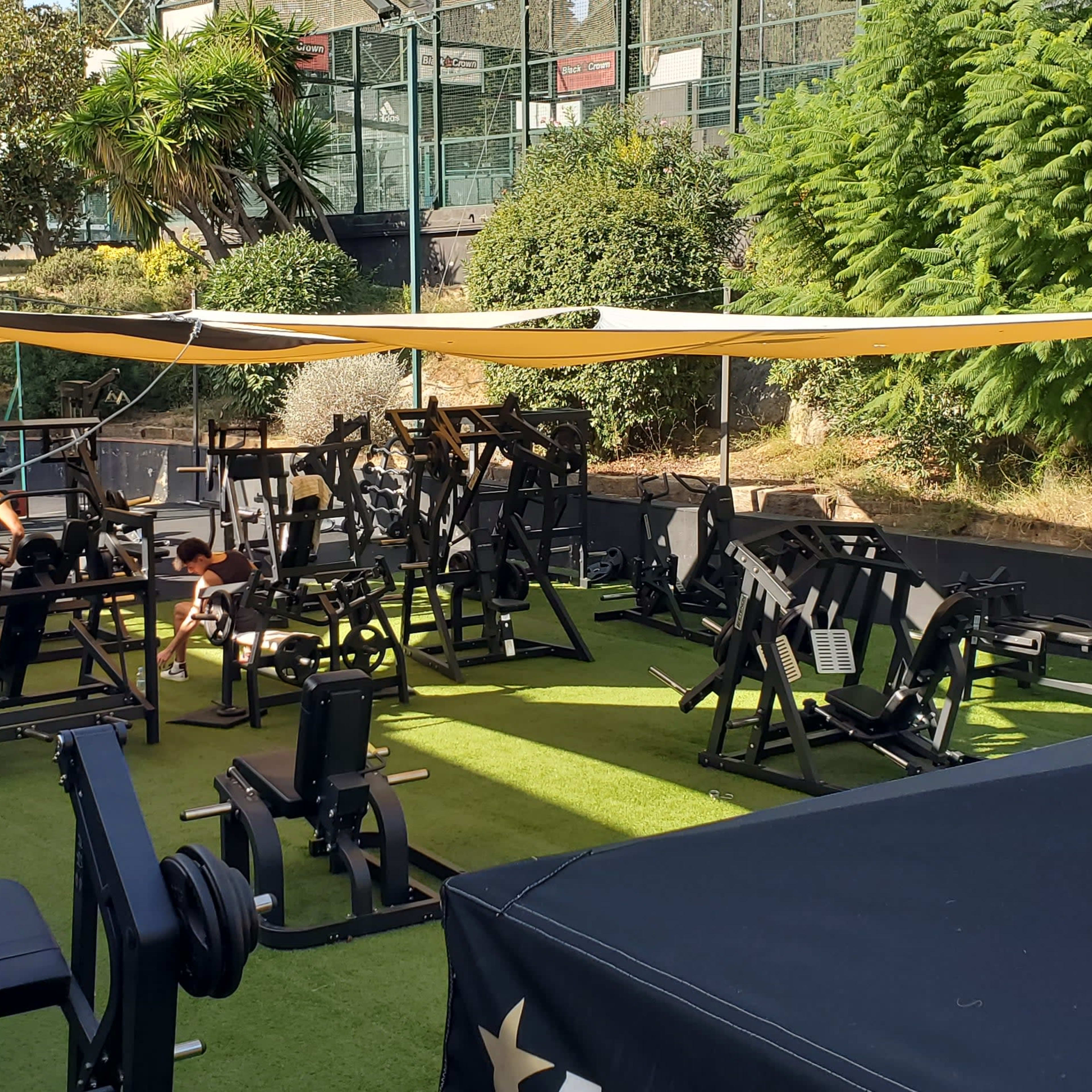 A partially shaded outdoor gym is equipped with various workout machines and weights, surrounded by greenery and a staircase in the background.