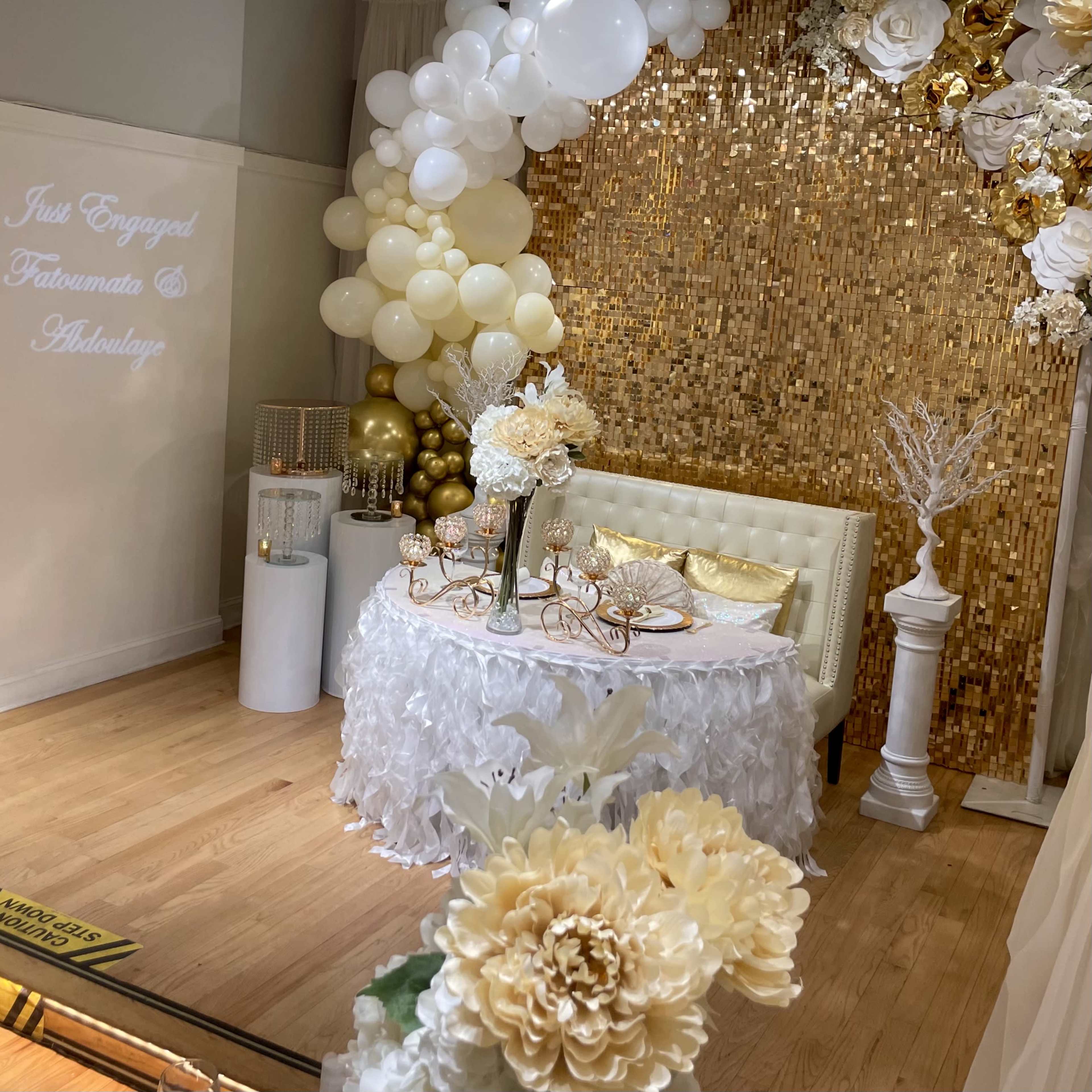 A decorated engagement celebration area featuring a white table with floral arrangements, a beige couch, and a backdrop of gold sequins and balloons.