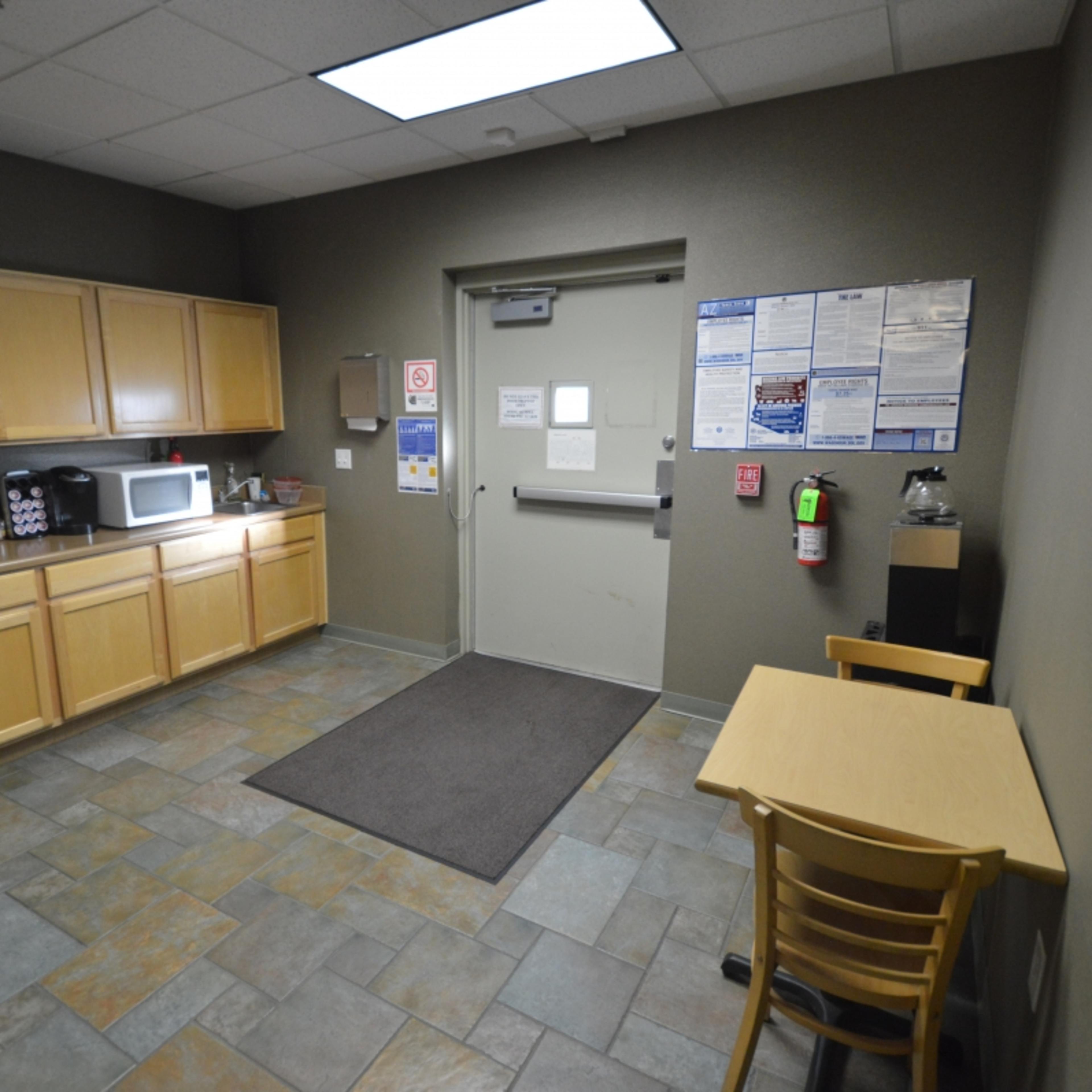 A break room with wooden cabinets, a microwave, a small table with a chair, and a door leading to the outside.