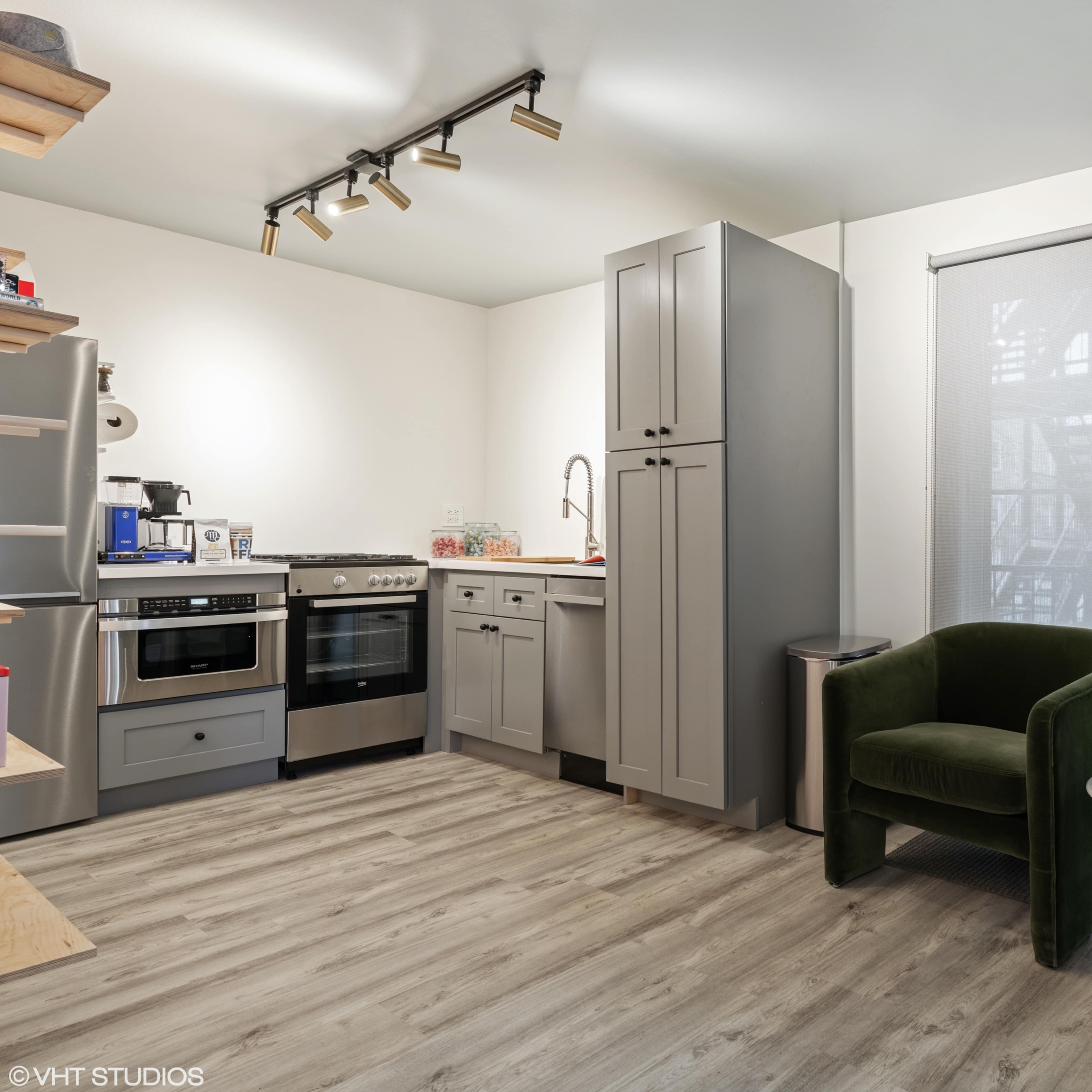 A compact kitchen area with modern appliances, gray cabinetry, and two green armchairs positioned beside a window.