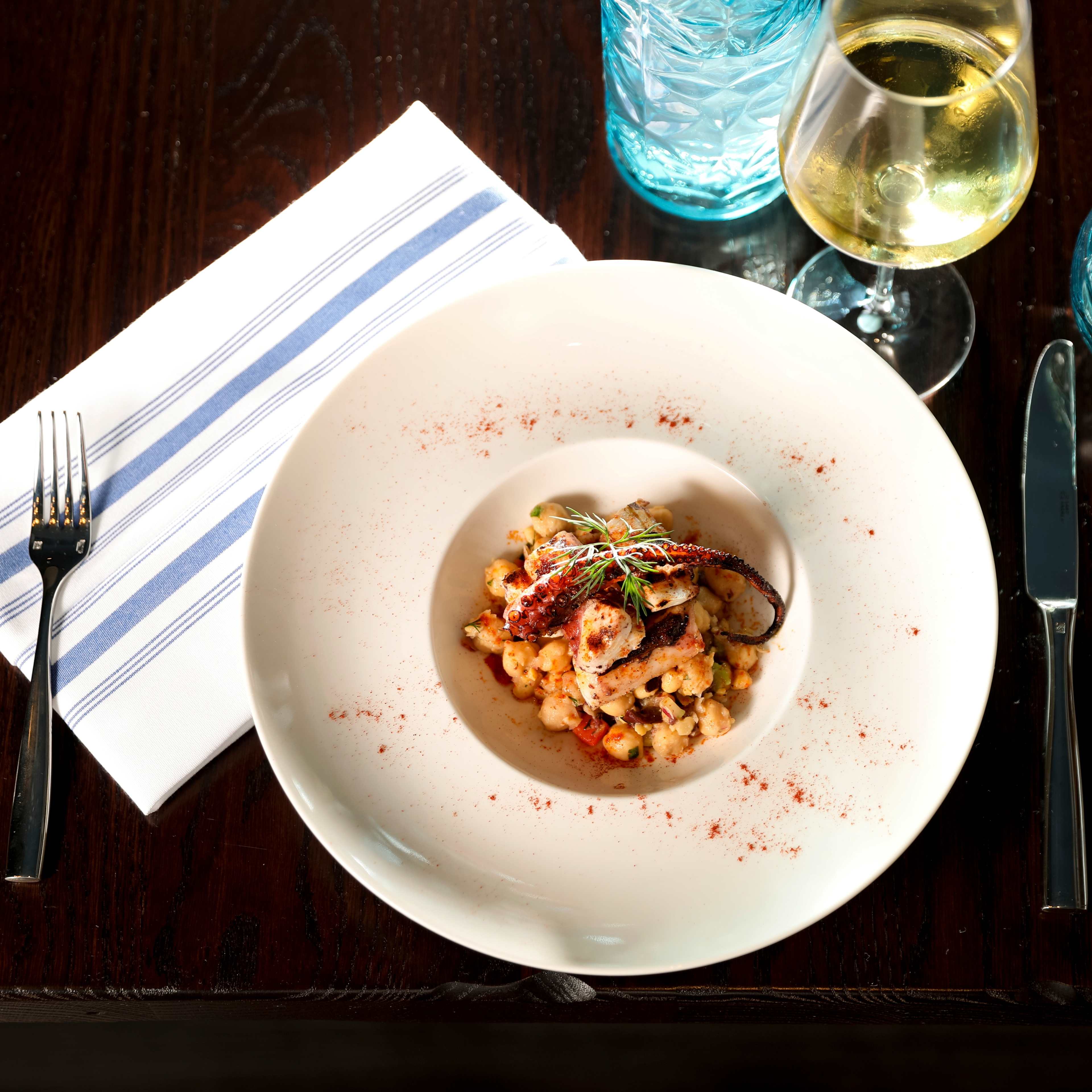 A plate of pasta with seafood is presented on a circular white dish, accompanied by a glass of white wine and a folded striped napkin.