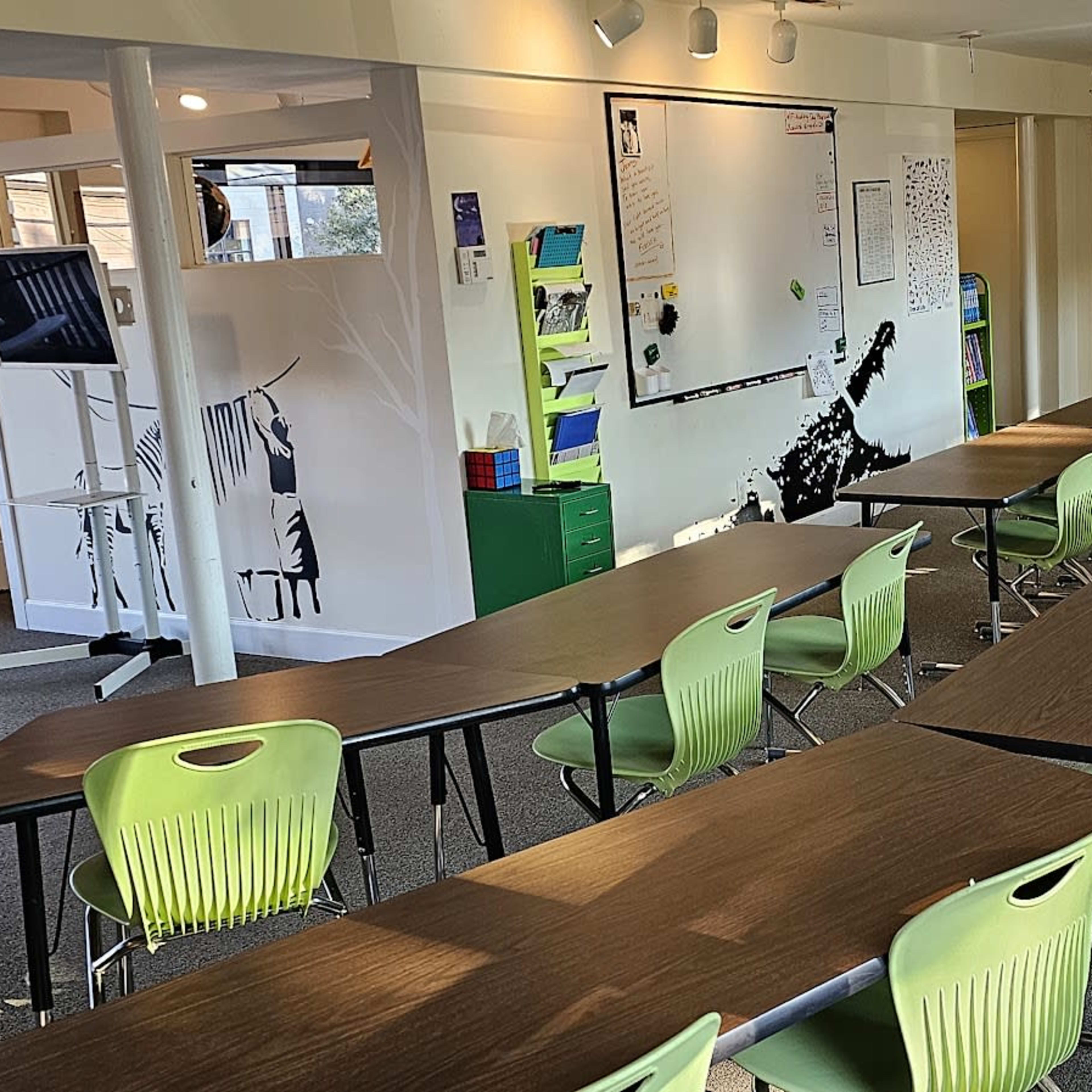 A classroom setup with several rows of tables and chairs, a whiteboard on the wall, and a monitor on a stand.