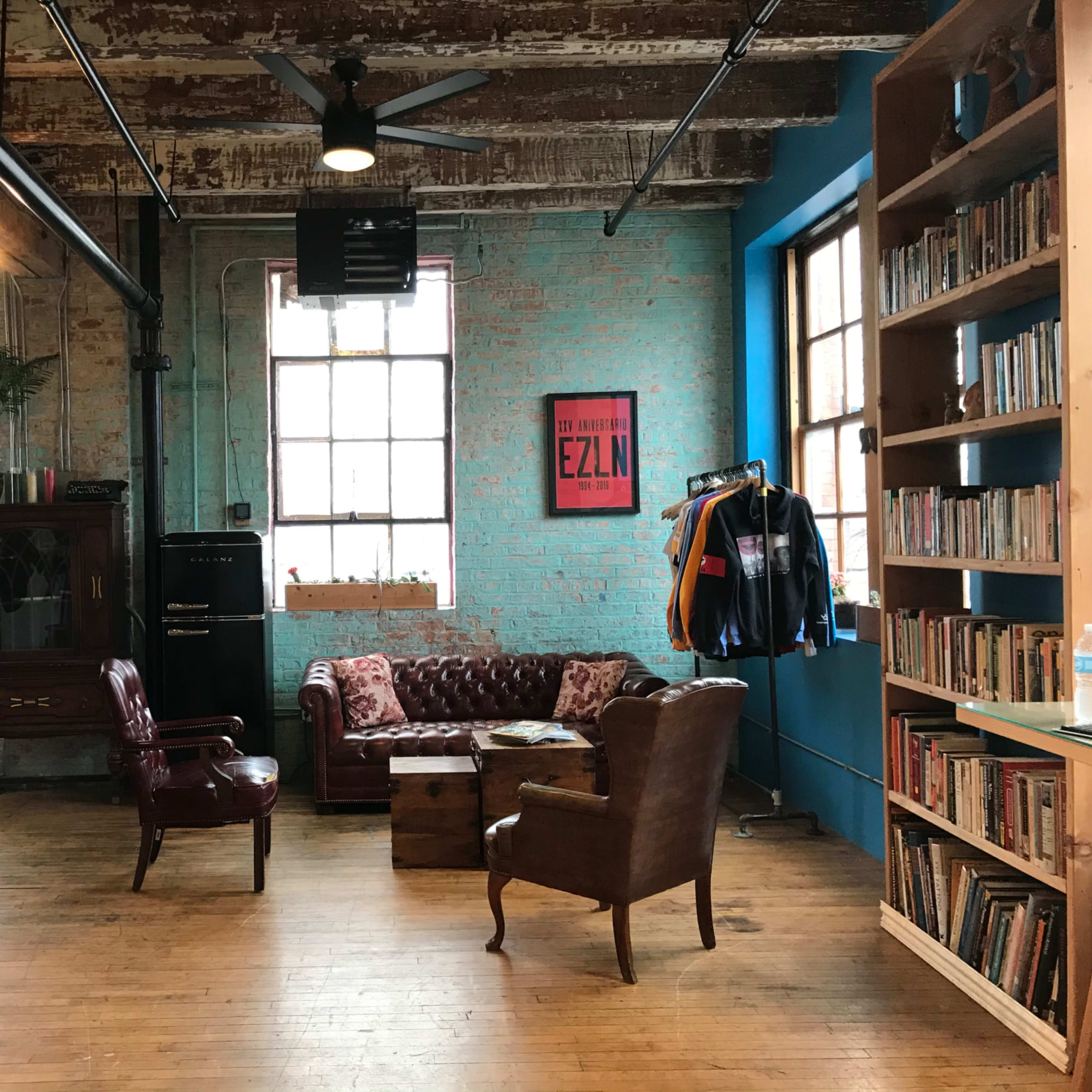 A spacious room with exposed brick walls, wooden floors, and a seating area featuring two armchairs and a coffee table, along with a bookshelf and a vintage cabinet.