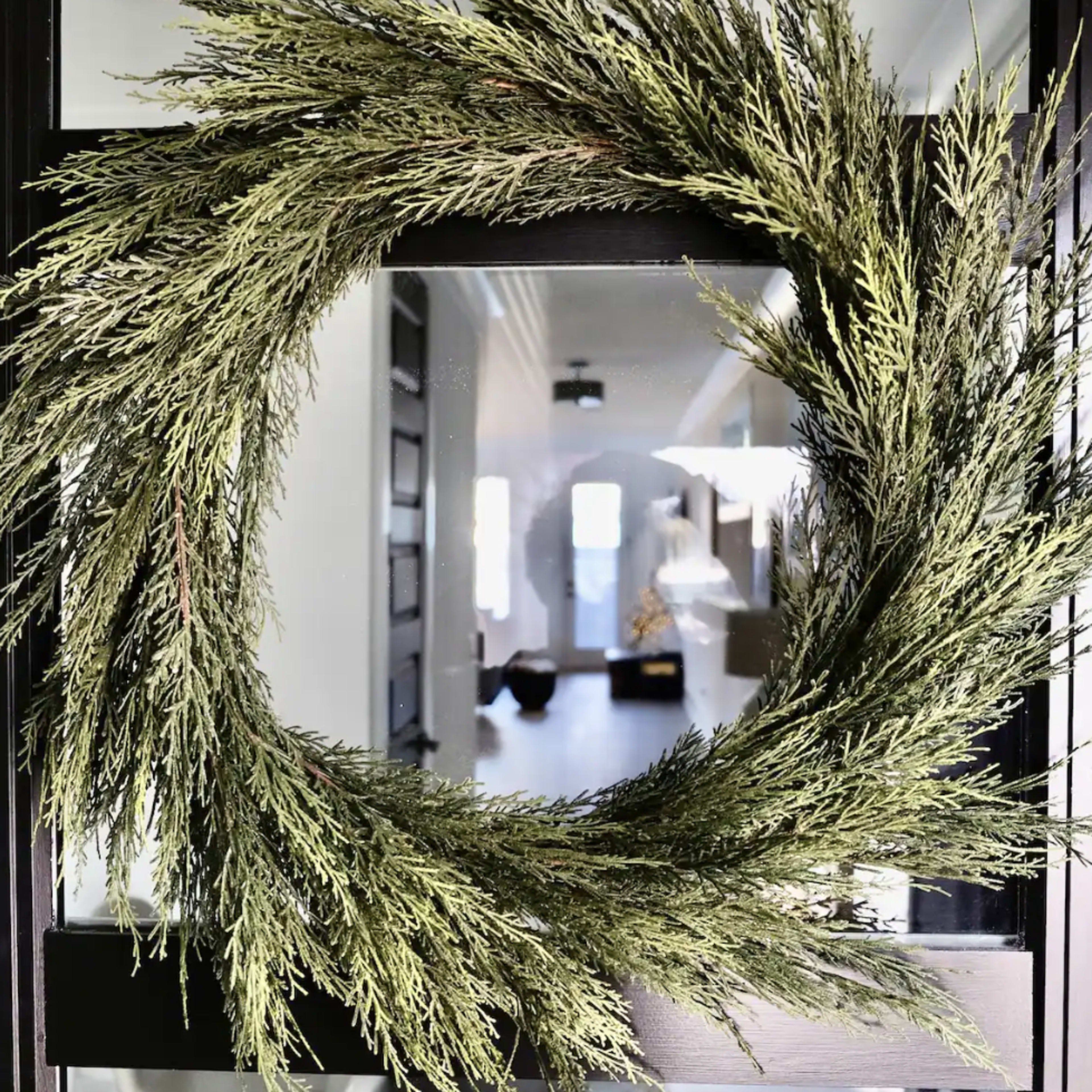 A green wreath made of evergreen branches hangs on a glass door, reflecting the interior of a well-lit room.