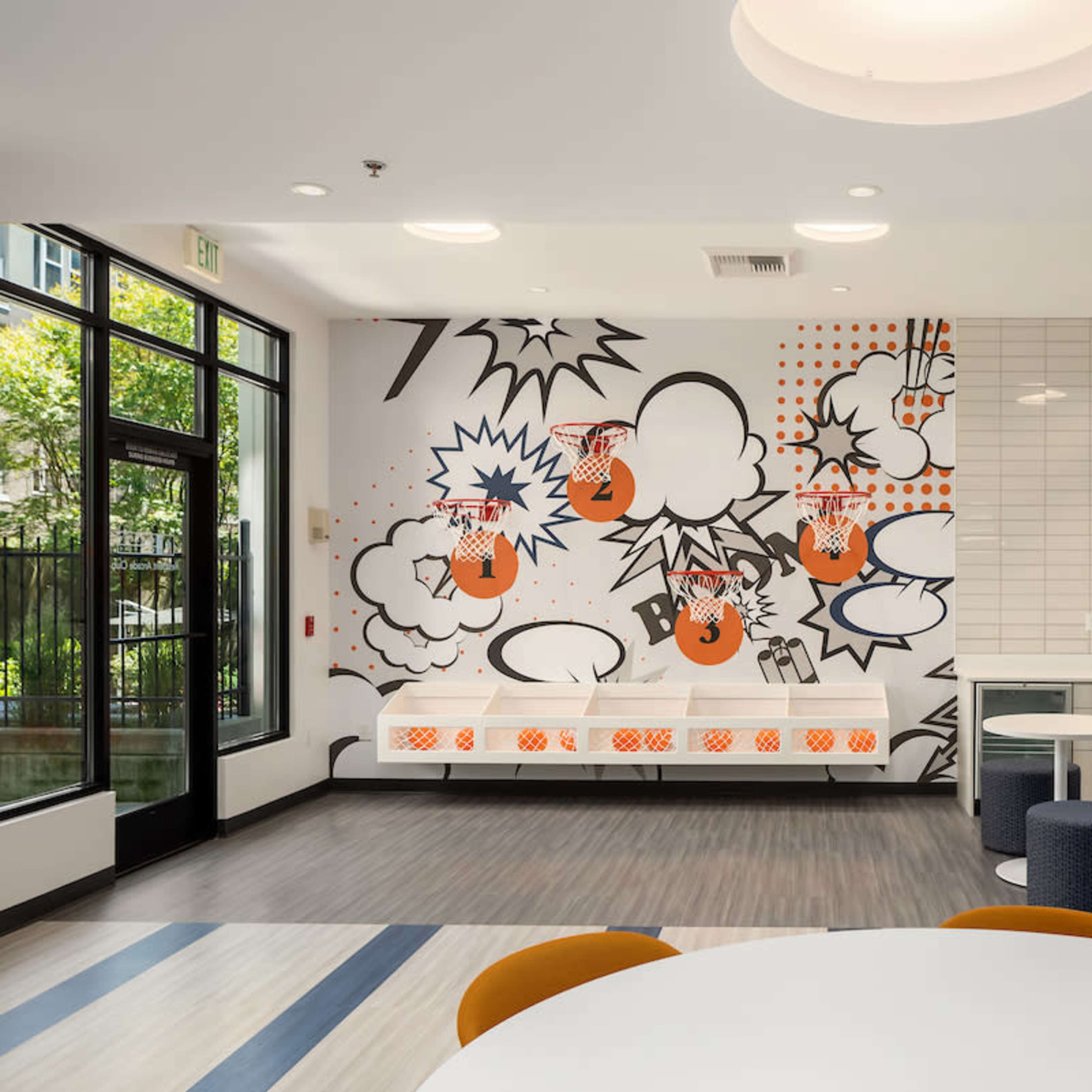 A modern communal space with a large graphic mural on one wall, a kitchenette area, and various seating options, including round tables and stools.