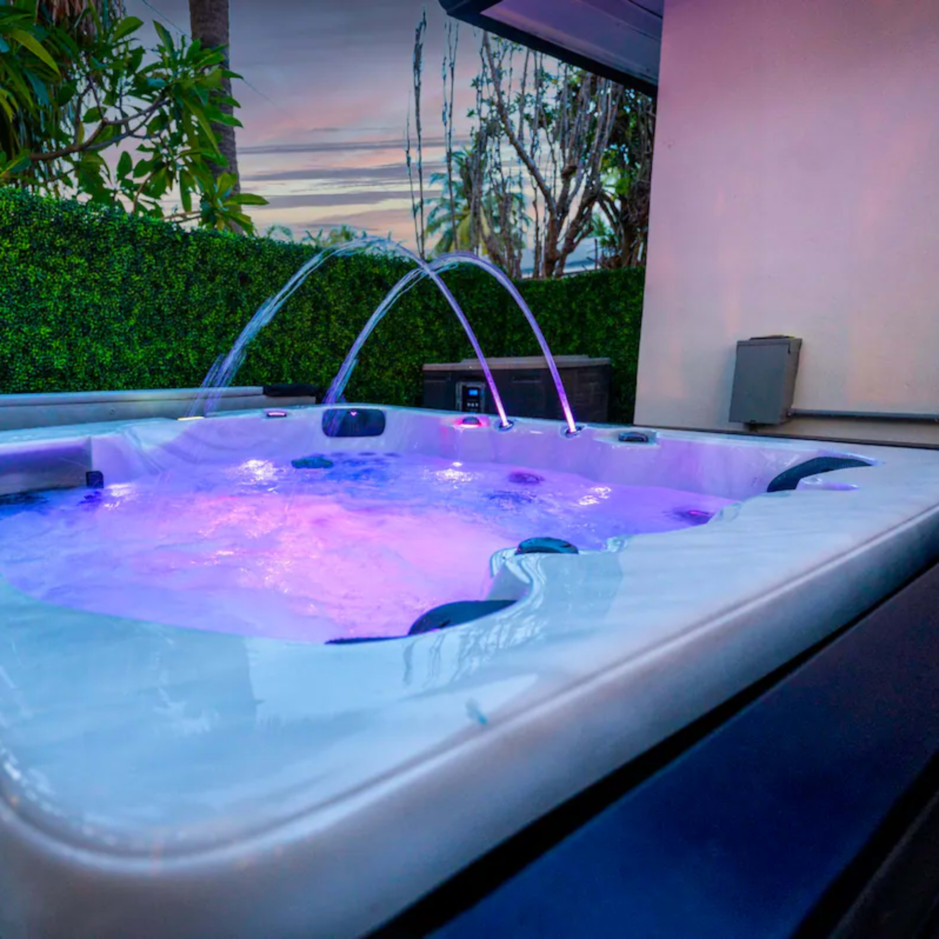 A hot tub with colorful lighting is surrounded by greenery and features water jets that spray upward.