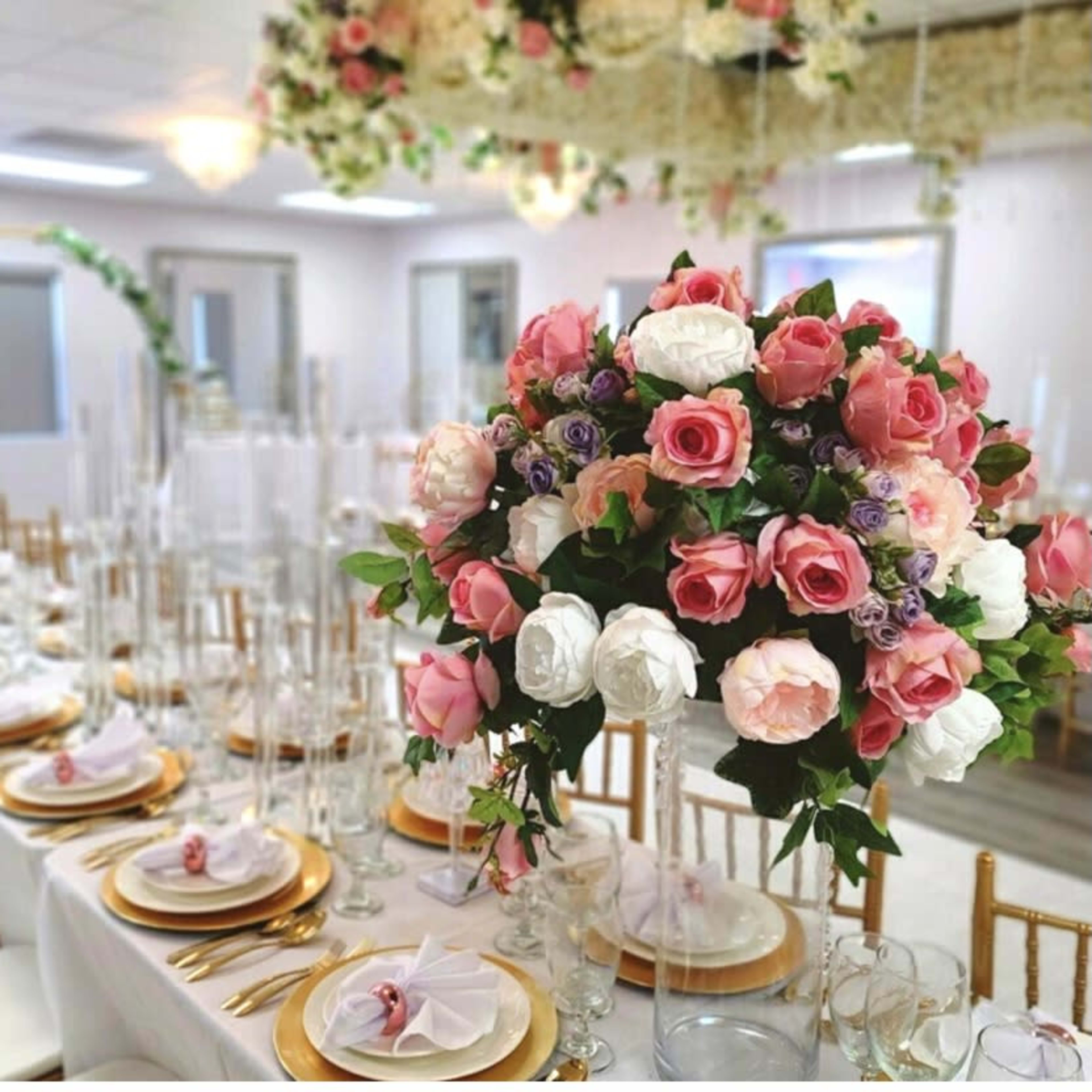 A banquet table is elegantly set with gold-rimmed plates, clear glassware, and a large floral centerpiece featuring pink and white roses.