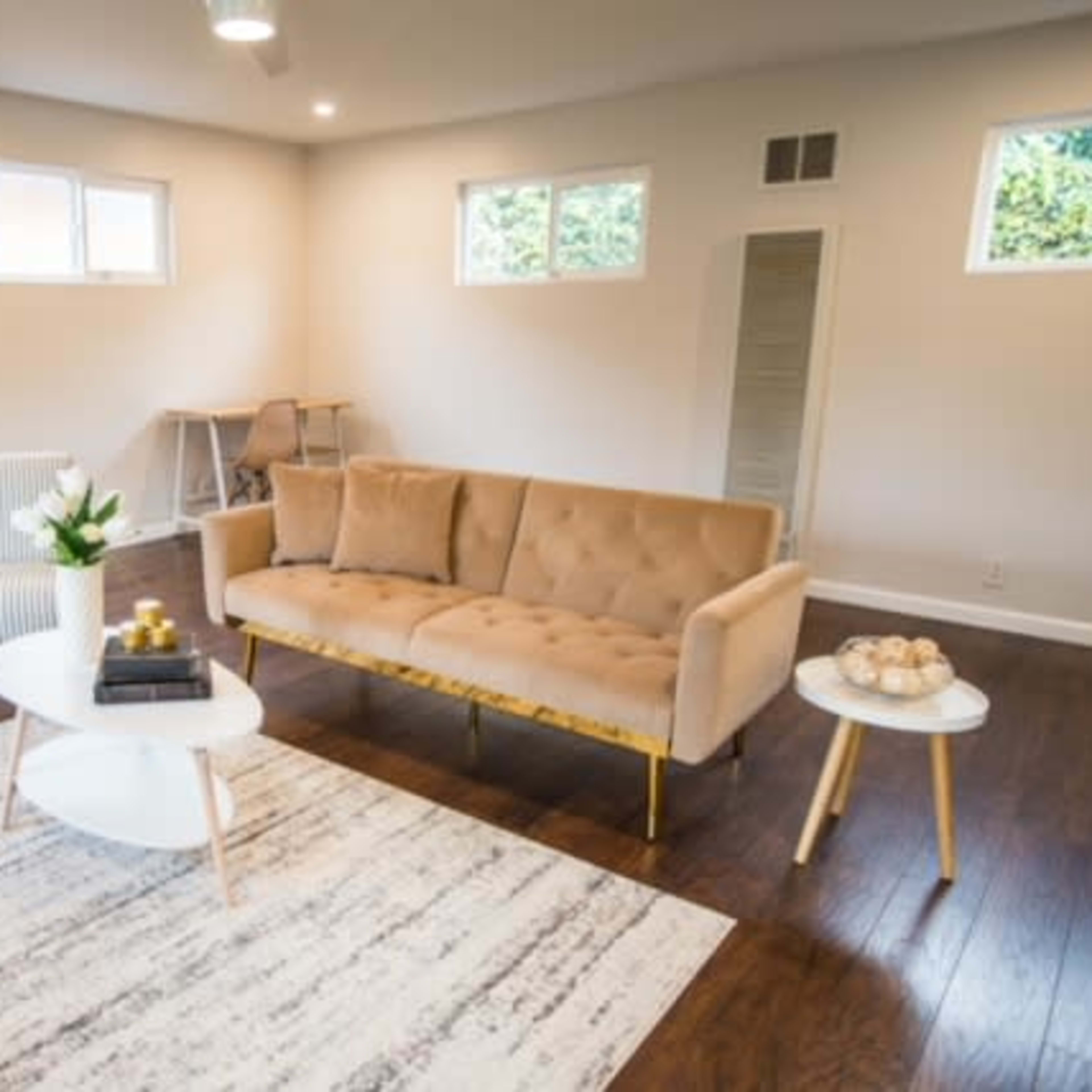 A modern living room with a beige tufted sofa, a small round coffee table, a patterned area rug, and large windows allowing natural light.