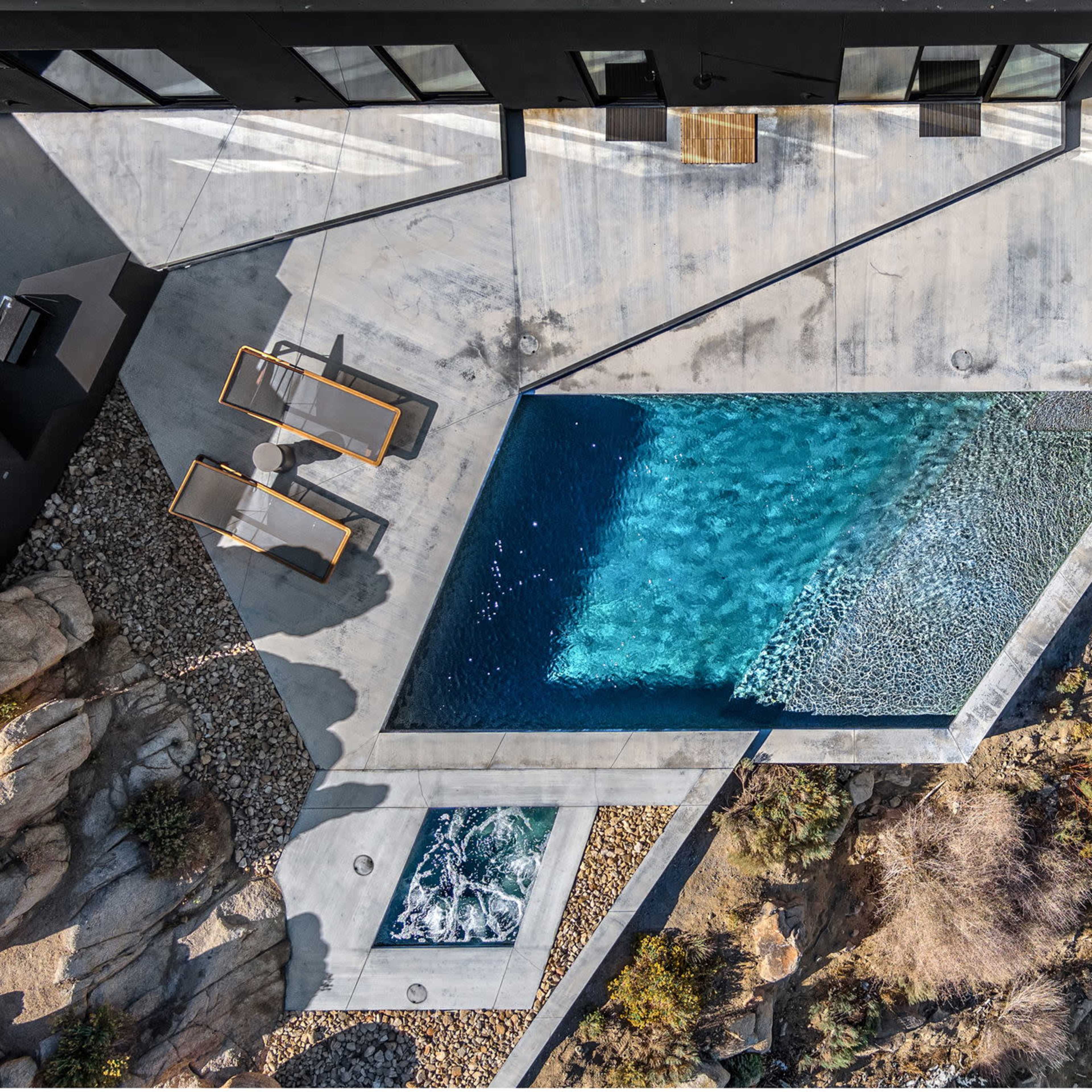 An aerial view shows a modern pool surrounded by concrete decking and rocky terrain, with two lounge chairs positioned nearby.