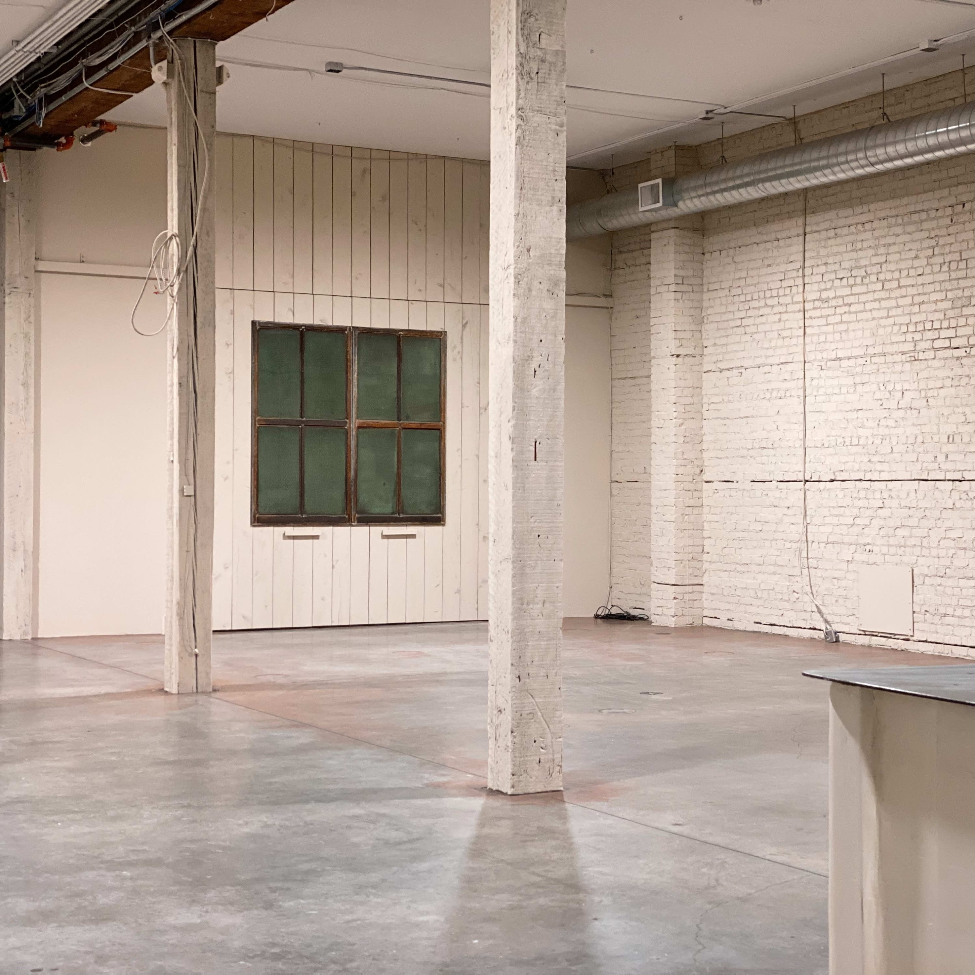 An empty industrial space with exposed concrete floors, white brick walls, wooden beams, and a window on one side.