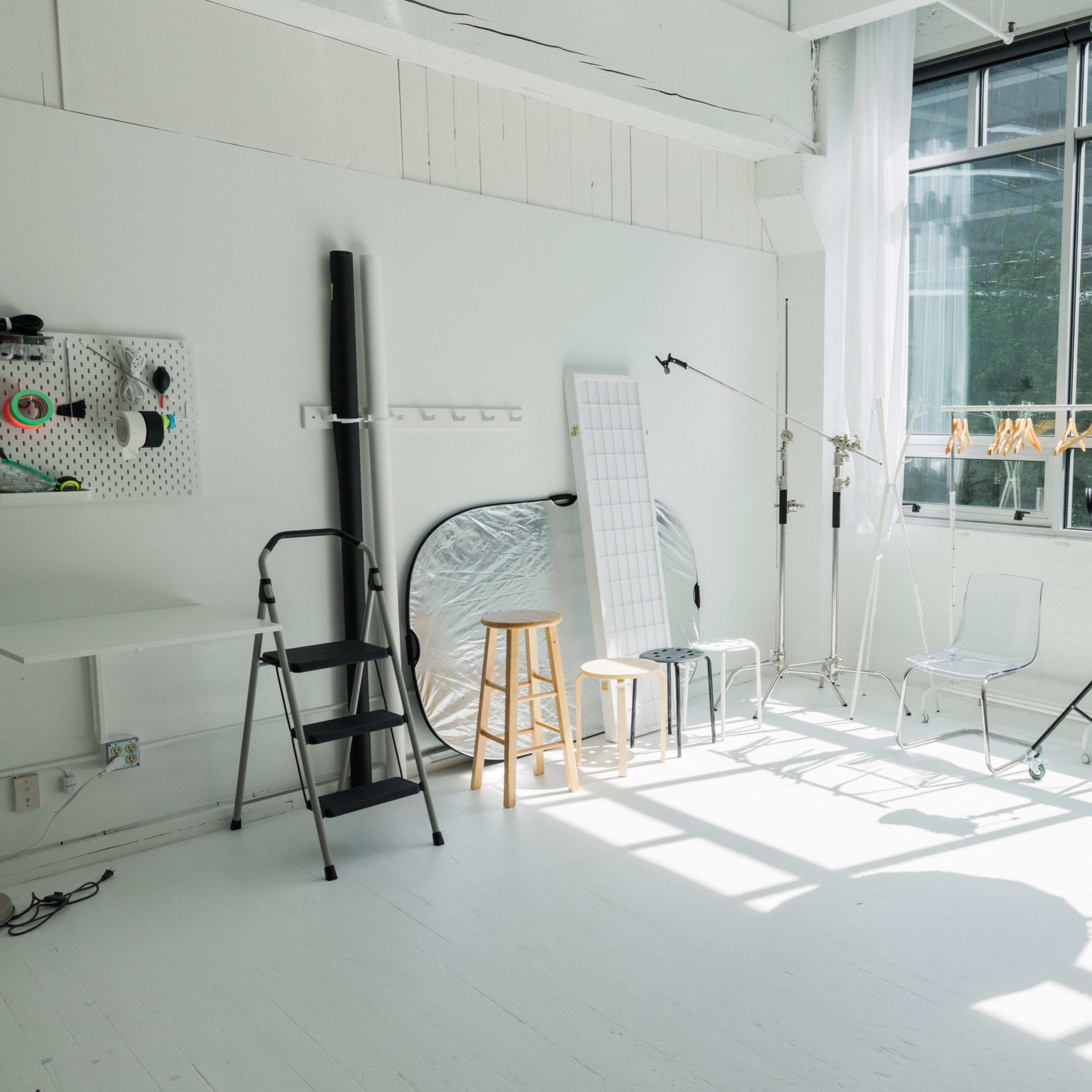 A well-lit photography studio featuring various equipment, including a large softbox, a reflector, and several chairs against a backdrop of large windows.